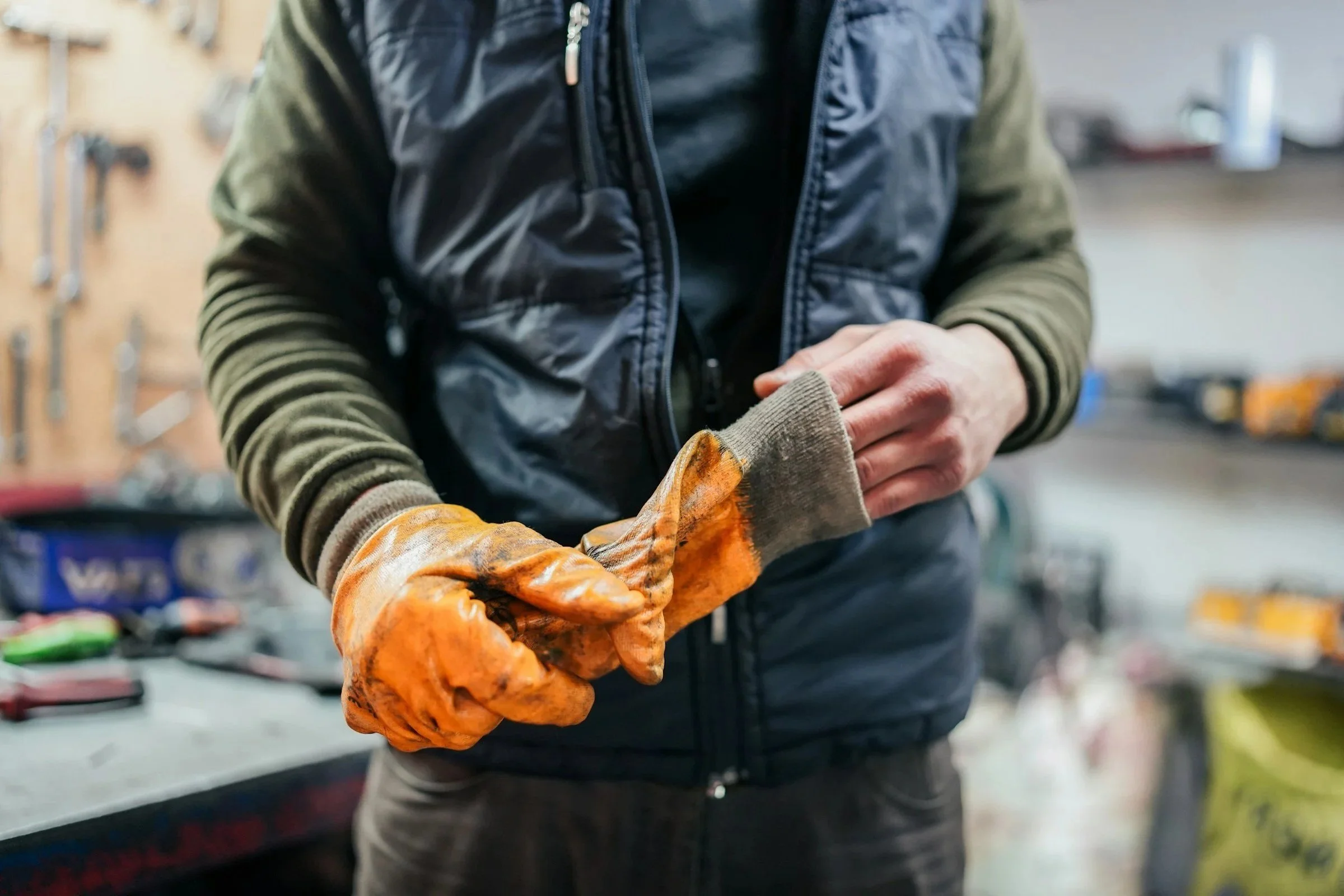Person wearing a black jacket and green hoodie working with orange work gloves in a workshop or hardware store.