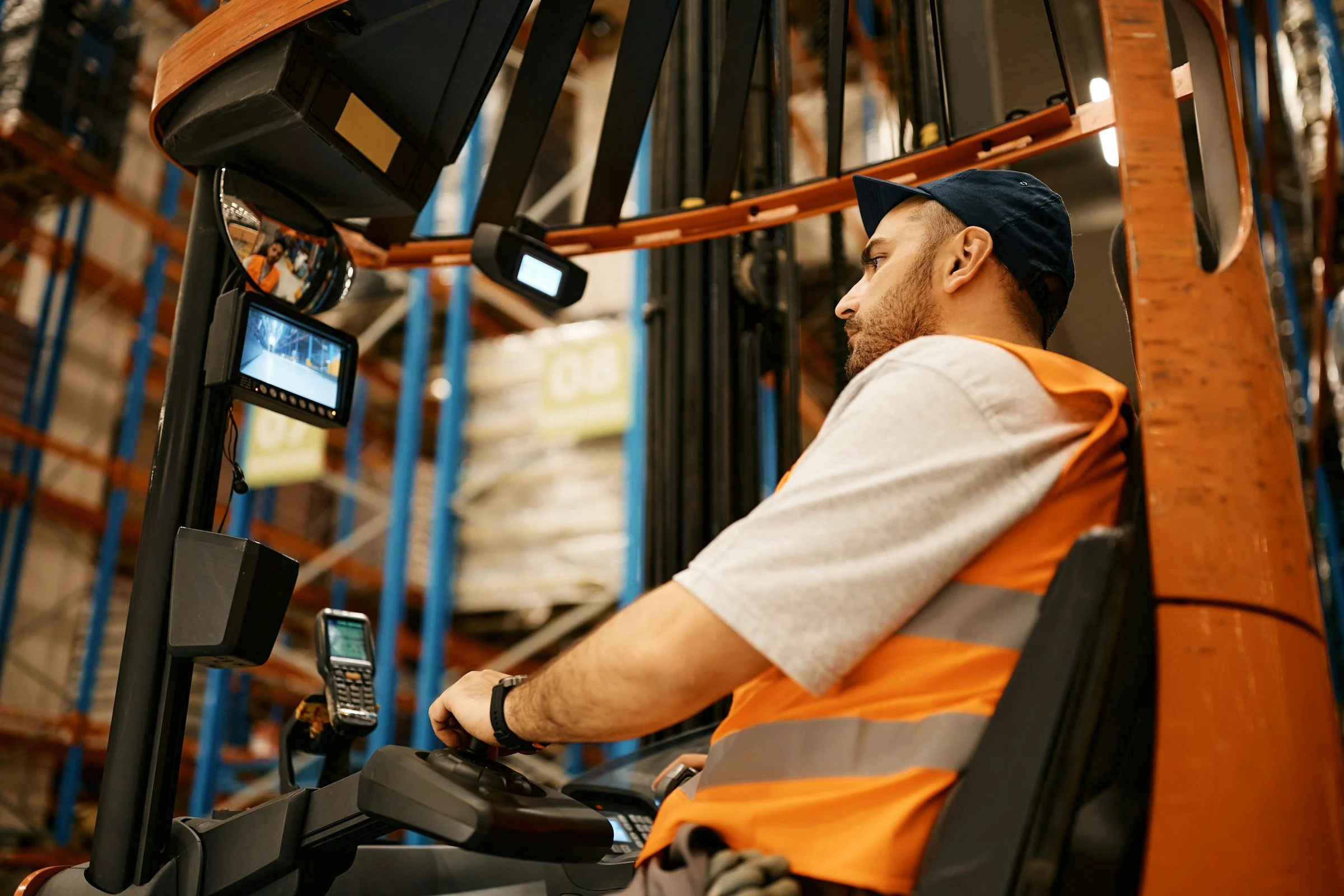 A warehouse worker wearing a navy baseball cap and an orange safety vest operates a forklift amidst warehouse shelves.
