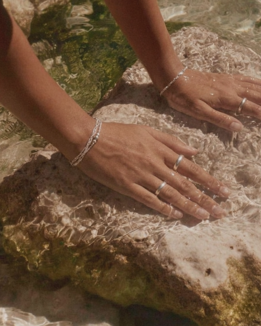 Hands with rings and bracelets resting on a wet rock in shallow water.