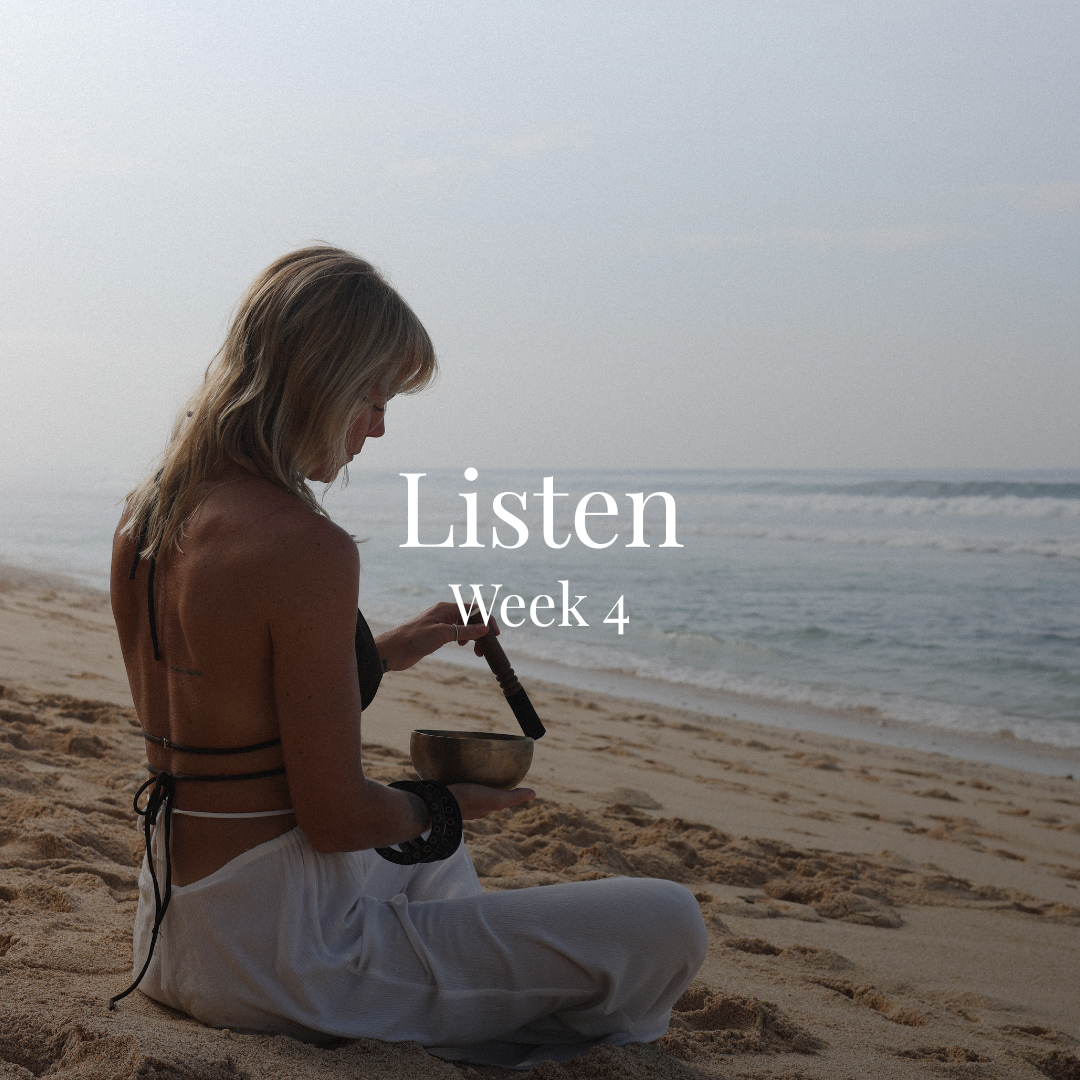 A woman sitting cross-legged on a sandy beach, playing a singing bowl, with the ocean and cloudy sky in the background, during daytime.