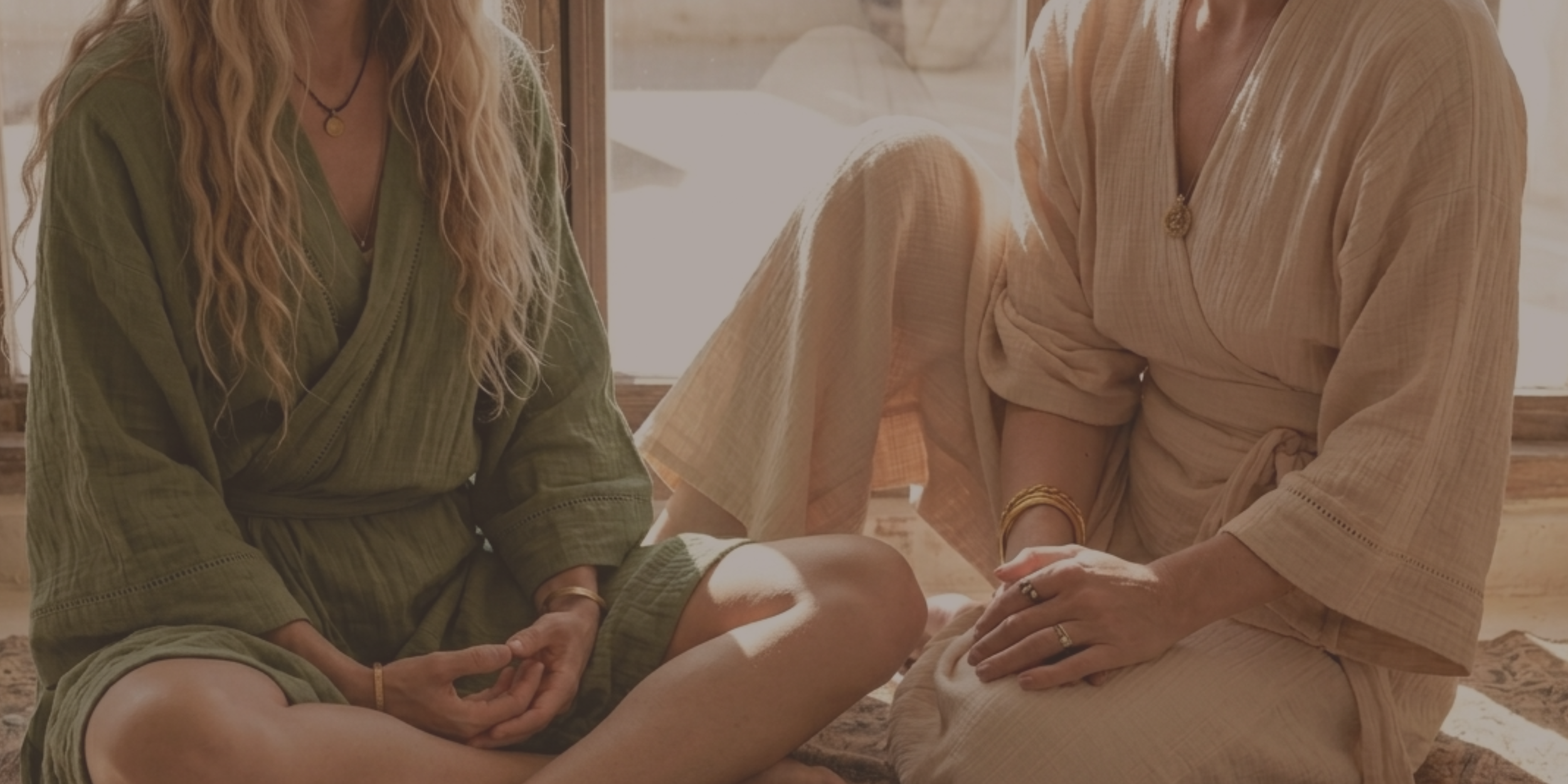 Two women sit close together on a wooden floor, holding hands, with sunlight streaming through nearby windows, creating a warm, intimate atmosphere.