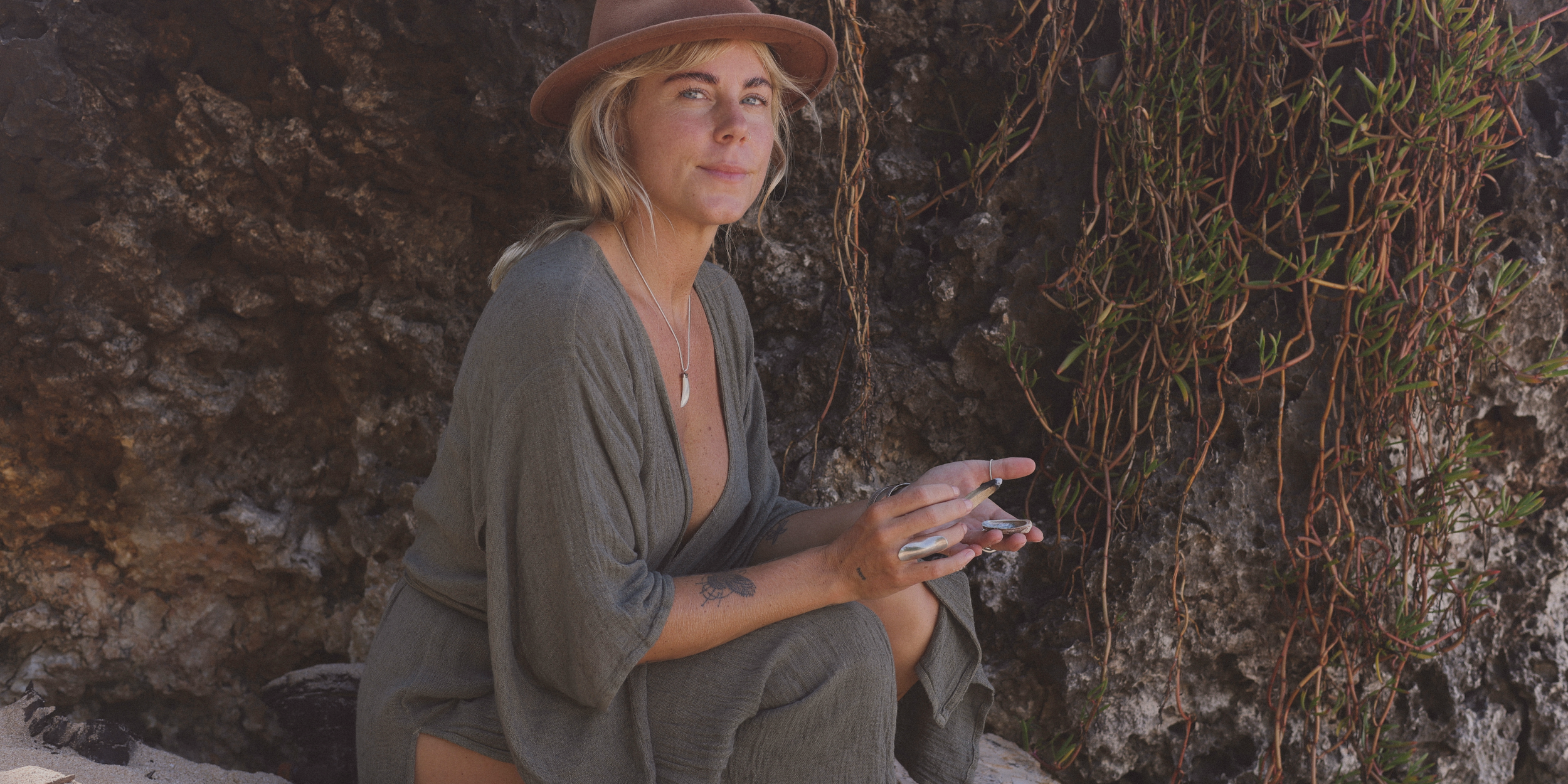 A young woman with blonde hair and tattoos, wearing a brown hat and gray clothing, sitting against a rocky wall with plants, holding a smartphone.