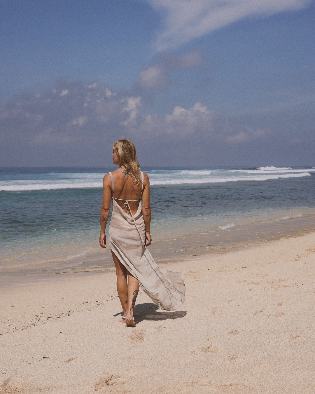 Woman with blonde hair walking on sandy beach near the ocean, wearing a long beige dress, with a partly cloudy sky overhead.