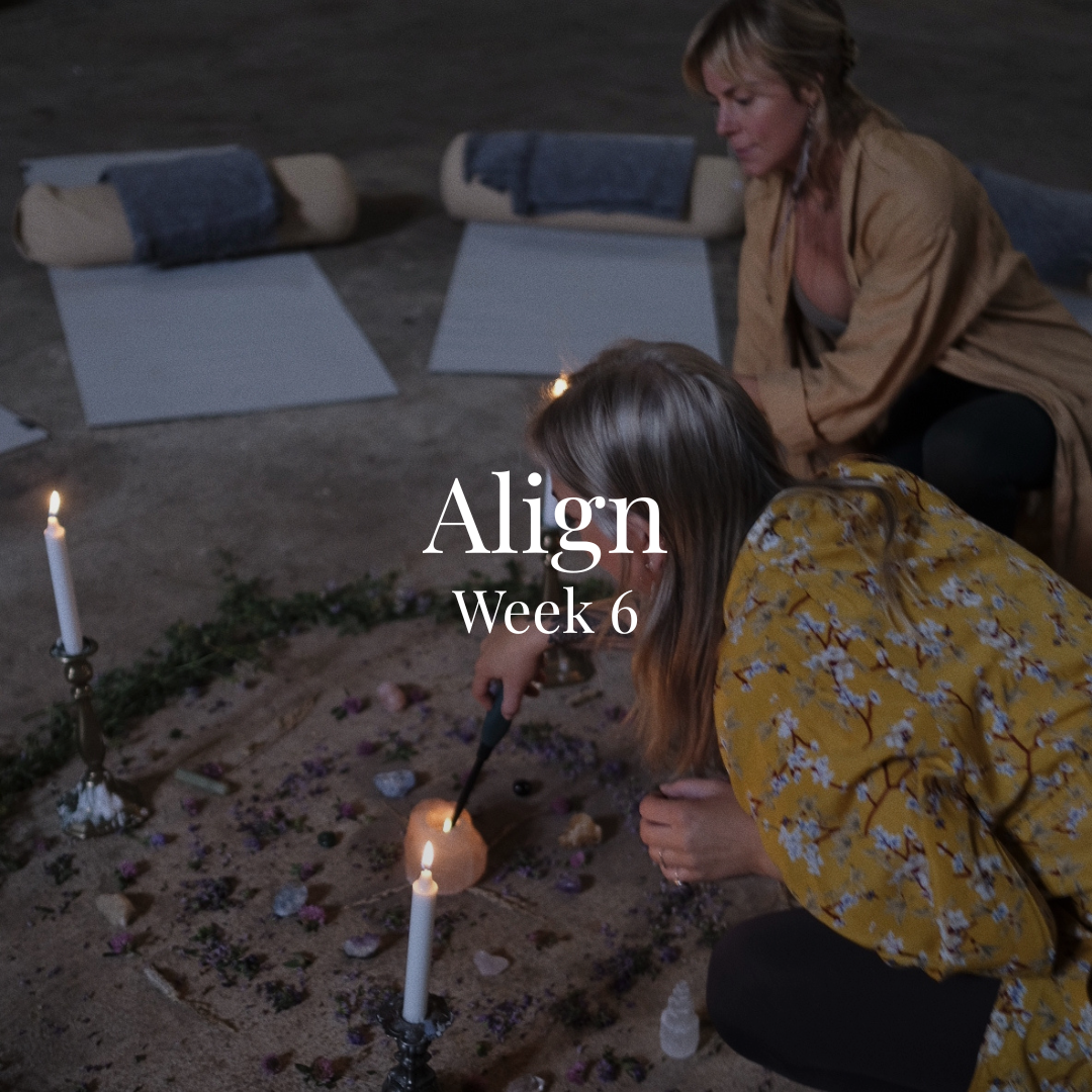 Two women performing a ritual on the ground with candles, crystals, and flowers, in a dimly lit space with yoga mats and rolled-up blankets in the background.