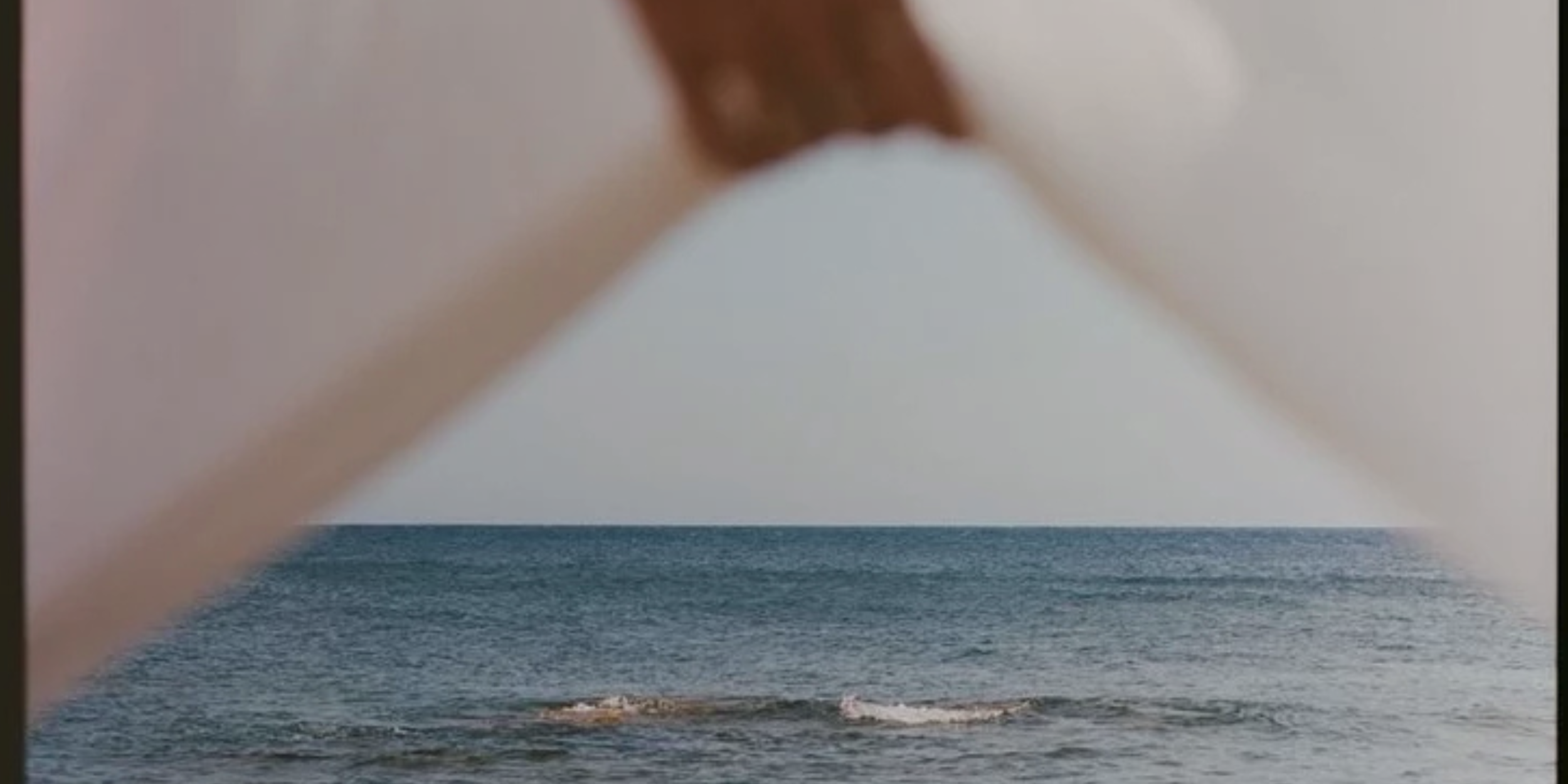 A close-up of a person's finger pinching the camera lens, with an ocean view scene in the background showing water and rocks under a cloudy sky.