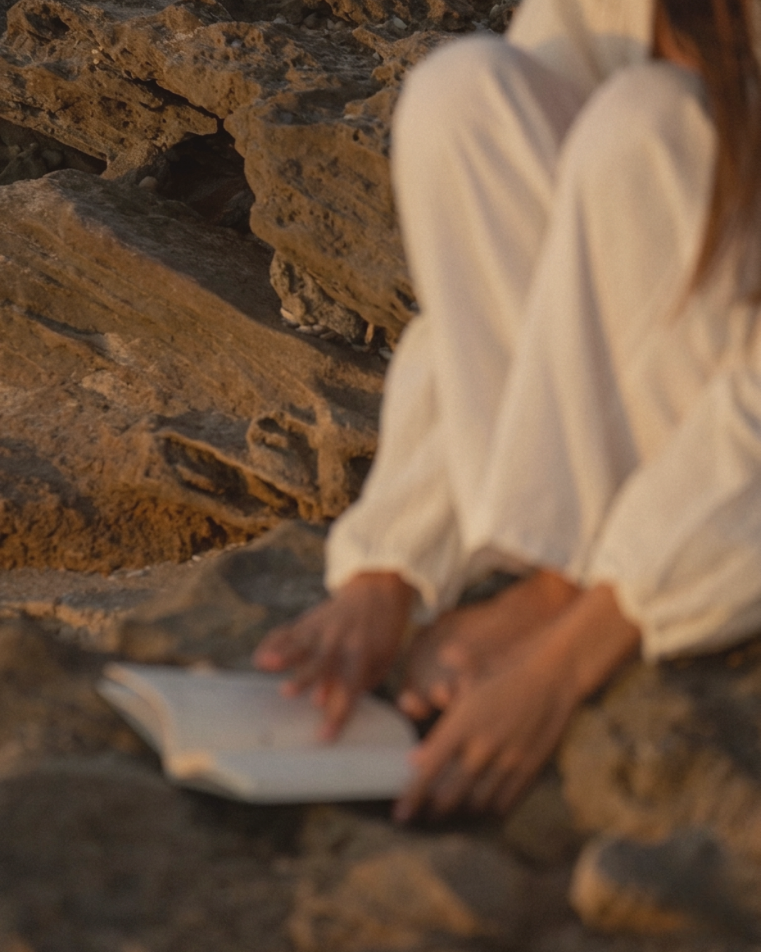 Person with long hair sitting on rocky ground, wearing a white long sleeve shirt, holding an open book.