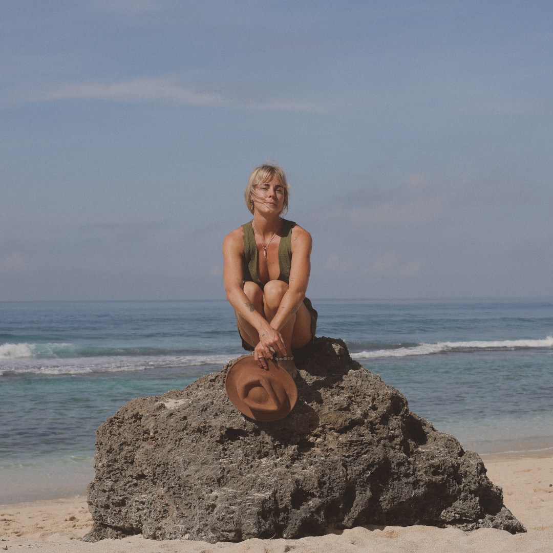 Woman sitting on a large rock at the beach, holding a brown hat, with ocean and blue sky in the background.