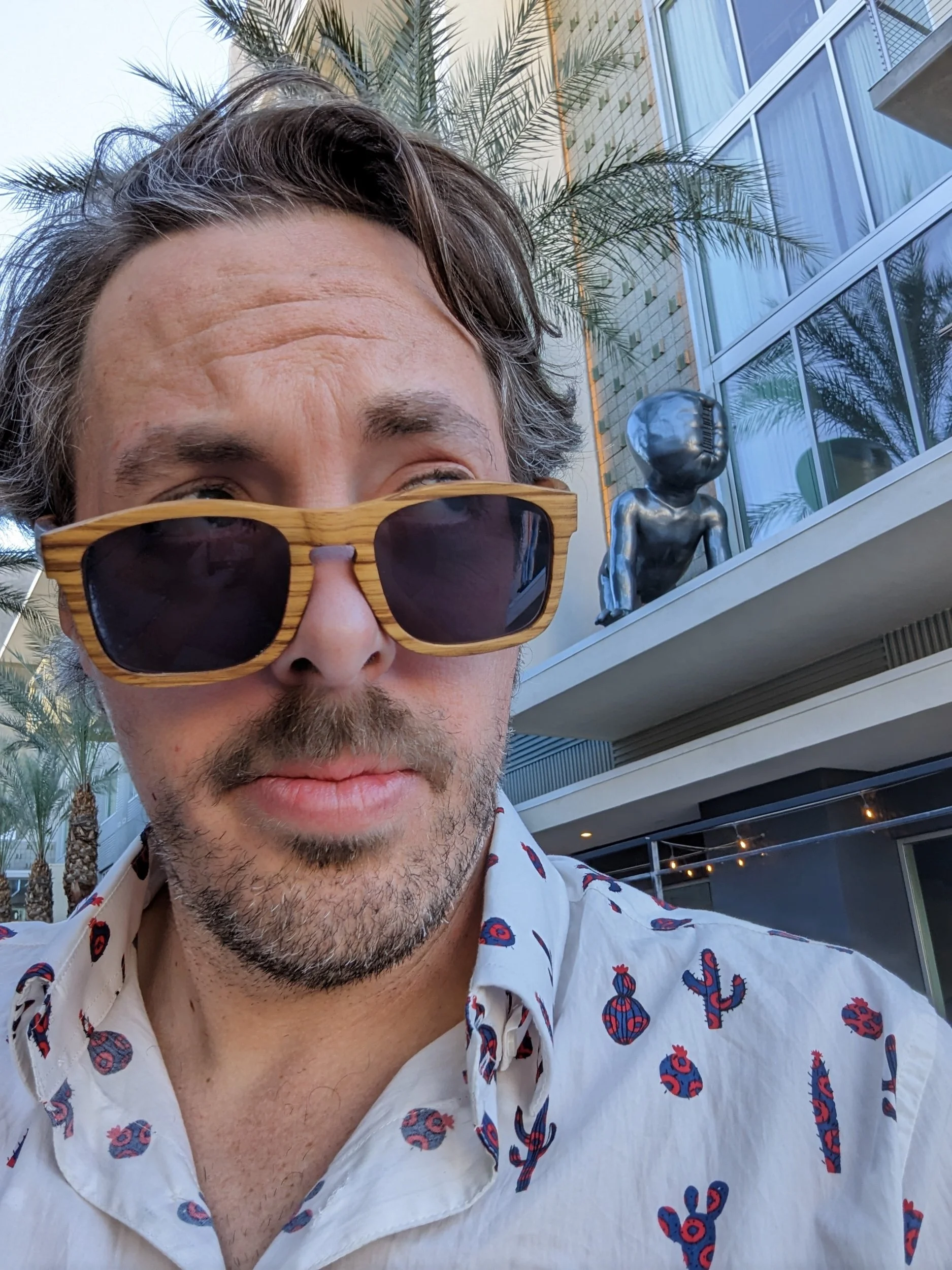 Close-up of a man with dark hair, facial hair, and light skin, wearing large wooden sunglasses and a white shirt with colorful cactus patterns, standing outdoors near a modern building with balconies, glass windows, palm trees, and string lights.