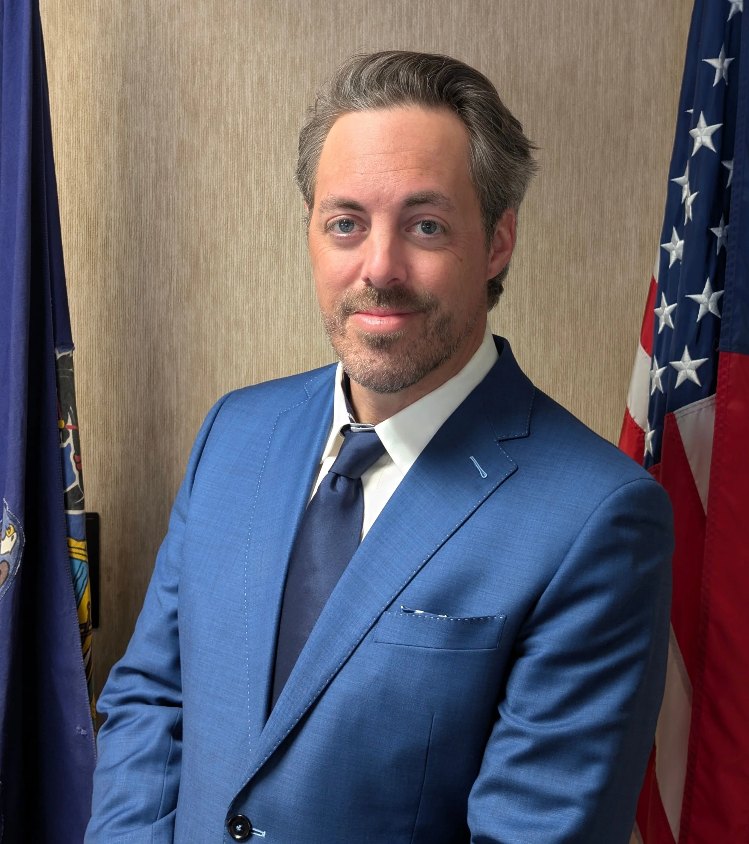 Close-up of a man with grey hair and a beard, wearing a blue suit and a black tie, standing in front of American flags.