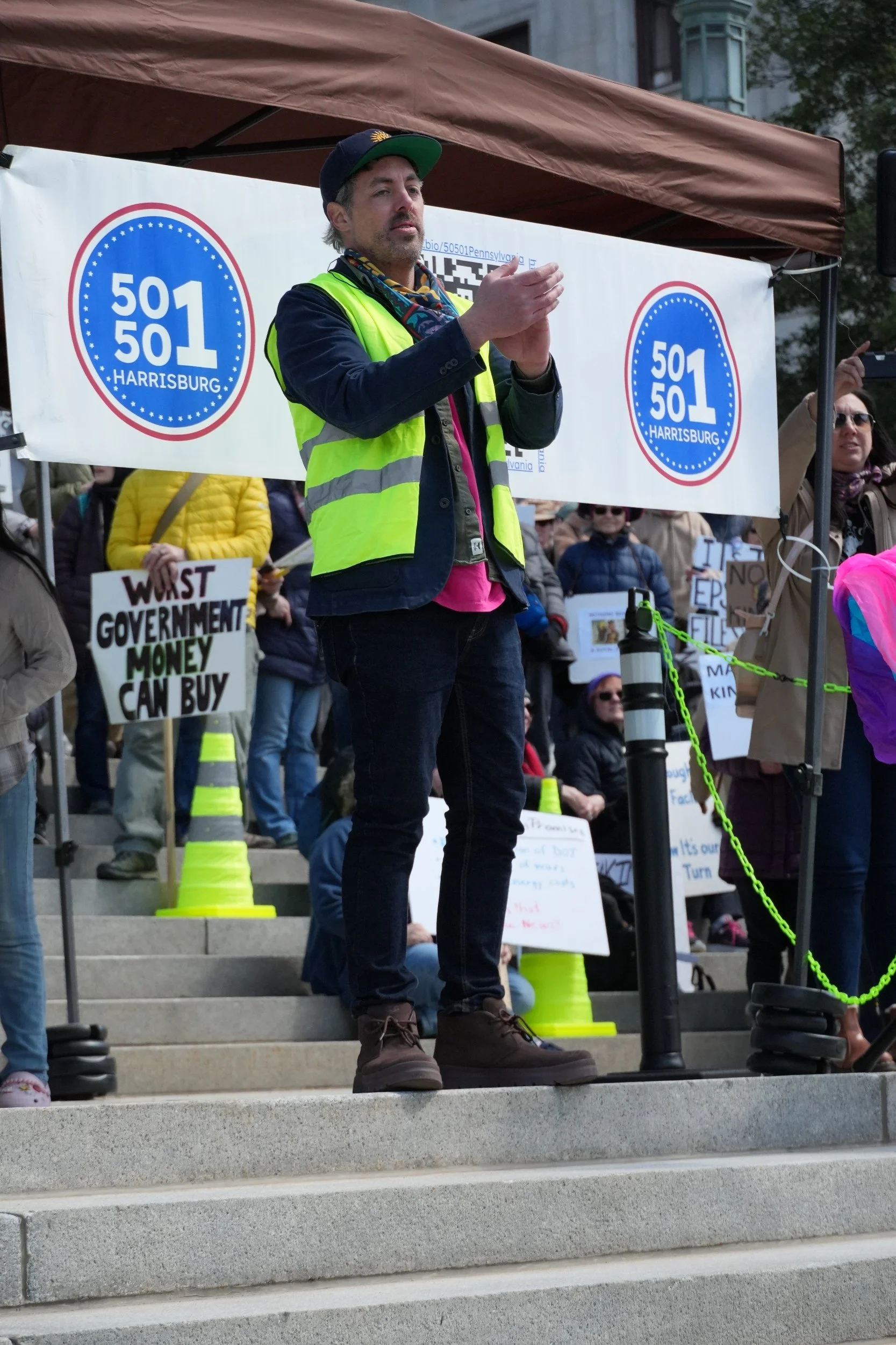 A man in a yellow safety vest standing on steps, addressing a crowd at a protest or rally. Behind him, a large banner displays the words '501 HARRISBURG' inside a blue circle with stars. Several signs are held by the crowd, one of which reads 'WASTED GOVERNMENT MONEY CAN BUY.' The event appears to be outdoors in an urban setting with a historic building or government building in the background.