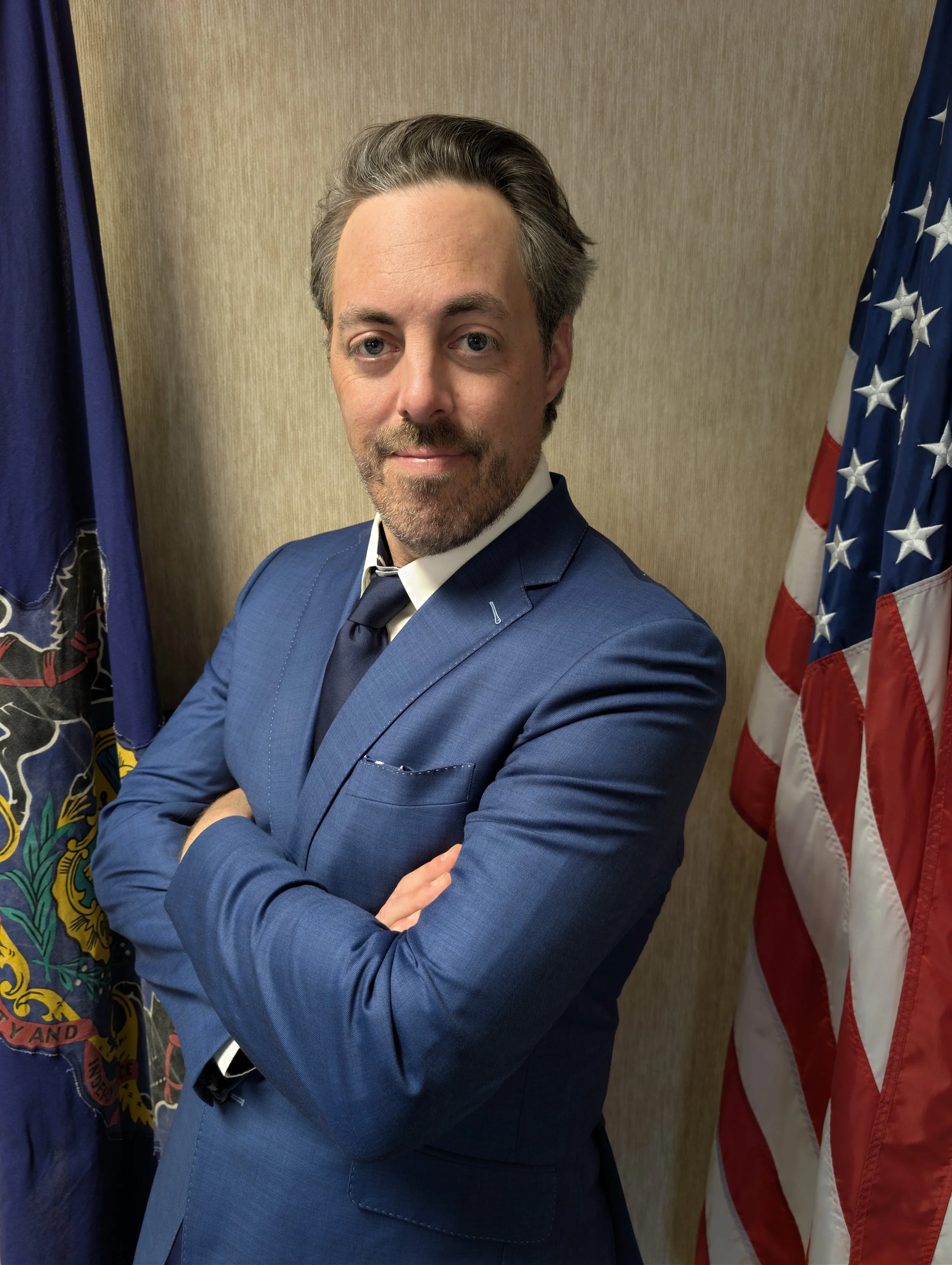 A man in a blue suit standing with arms crossed, flanked by an American flag and a state flag, in front of a beige wall.