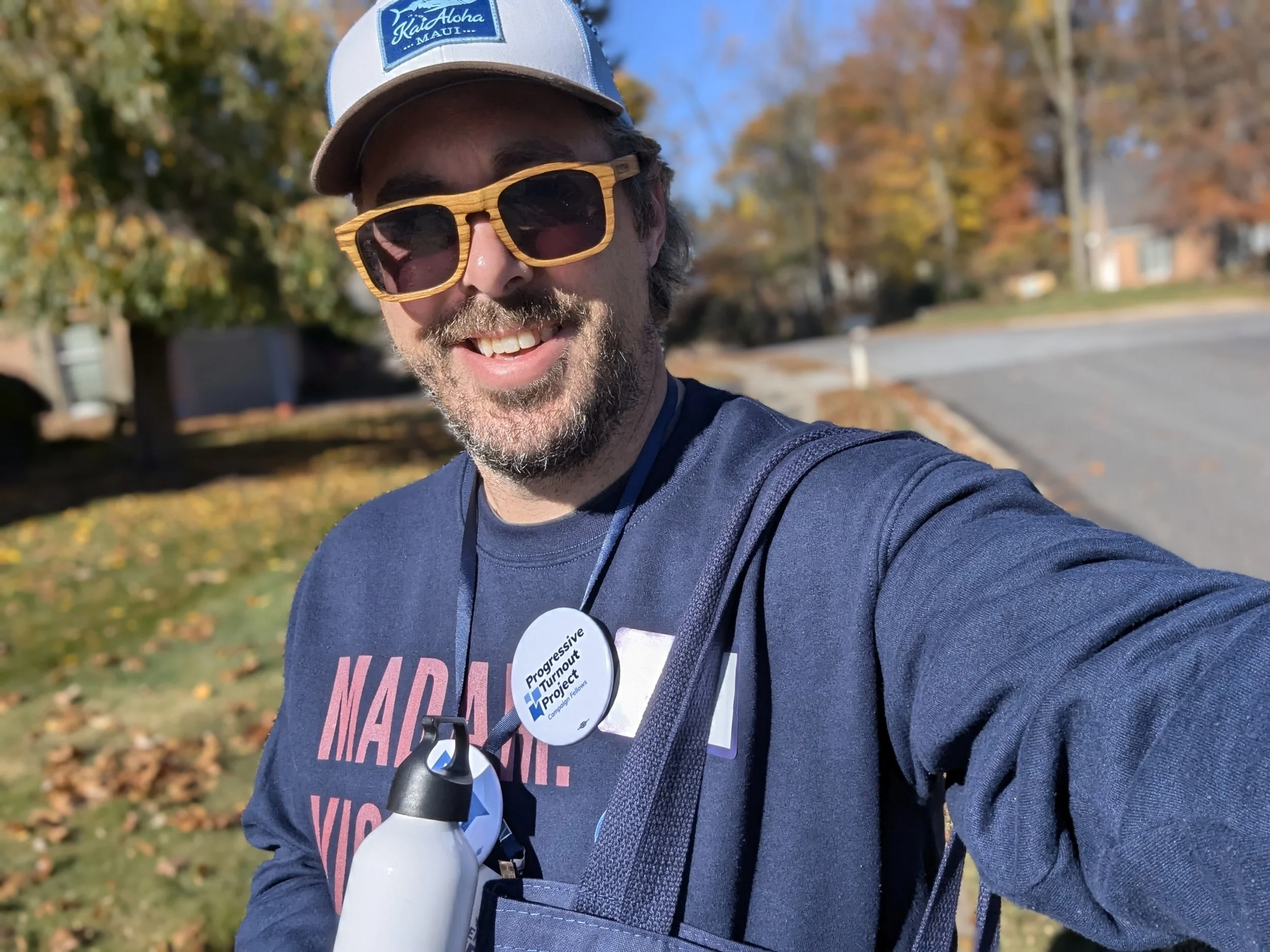 A man with curly hair and a beard wearing sunglasses, a baseball cap, a navy blue shirt, and event badges takes a selfie outdoors on a sunny fall day, with trees and a street in the background.
