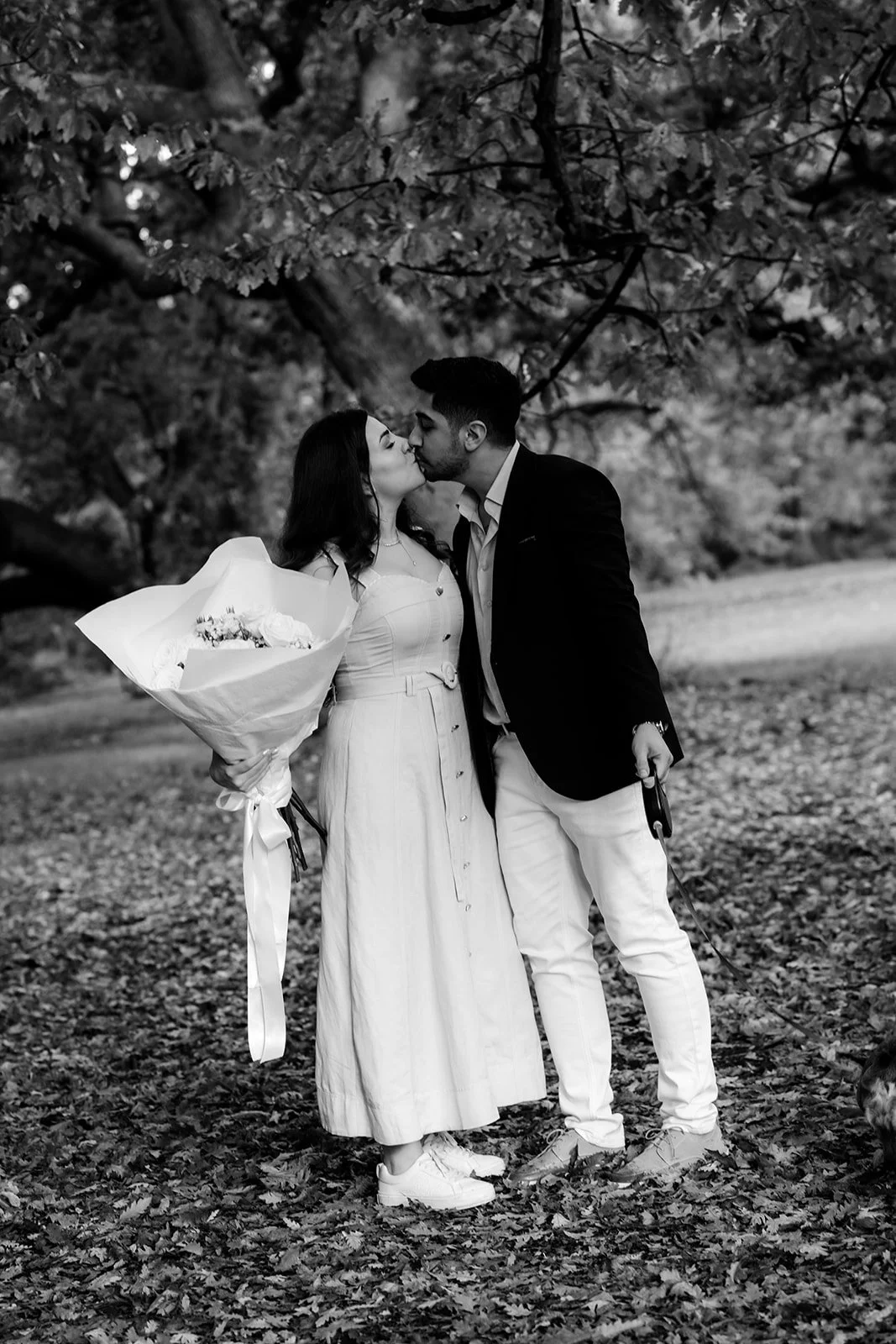 A black and white photo of a couple kissing outdoors, the woman holding a bouquet of flowers and wearing a dress, the man in a blazer and white pants, under a large tree.