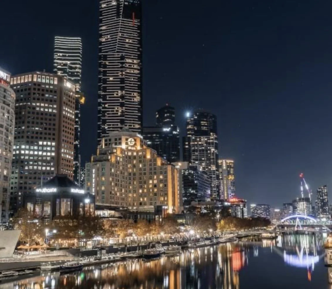 Night view of a city skyline with tall illuminated buildings and their reflections on the river below, including a bridge with bright lights.