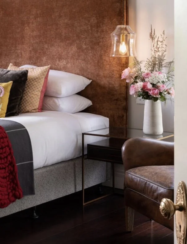 A cozy bedroom corner with a bed, pillows, a side table, a hanging light, and a large vase of pink and white flowers.