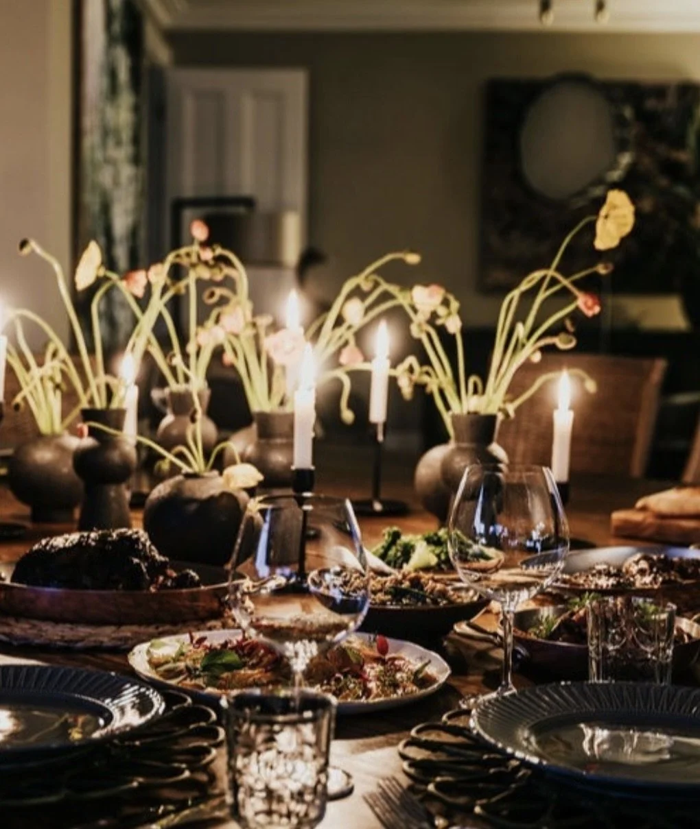 A dining table decorated with multiple vases of flowers, lit candles, and set with plates, glasses, and cutlery, in a dimly lit room.