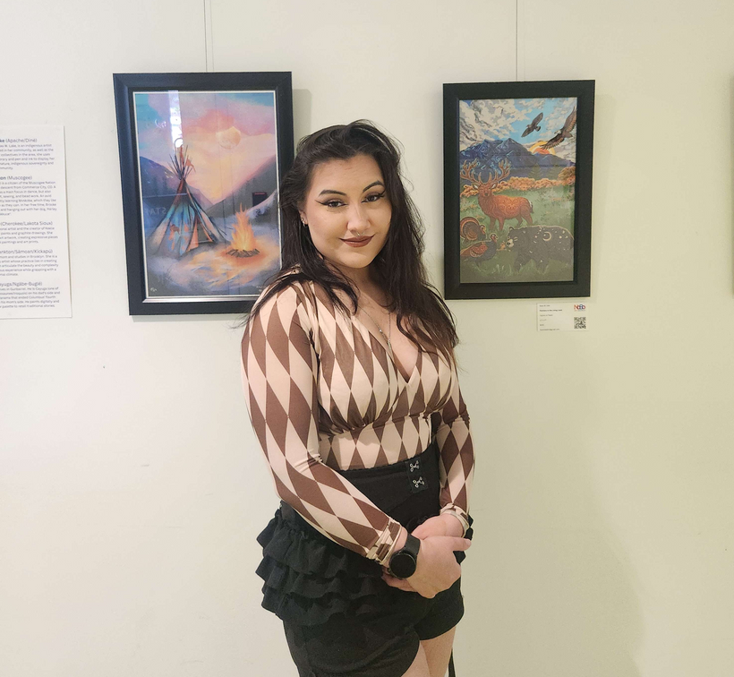 A woman standing in front of artwork in a gallery, with two framed paintings behind her, one depicting a tipi with a campfire and the other showing a mountain landscape with elk and birds.