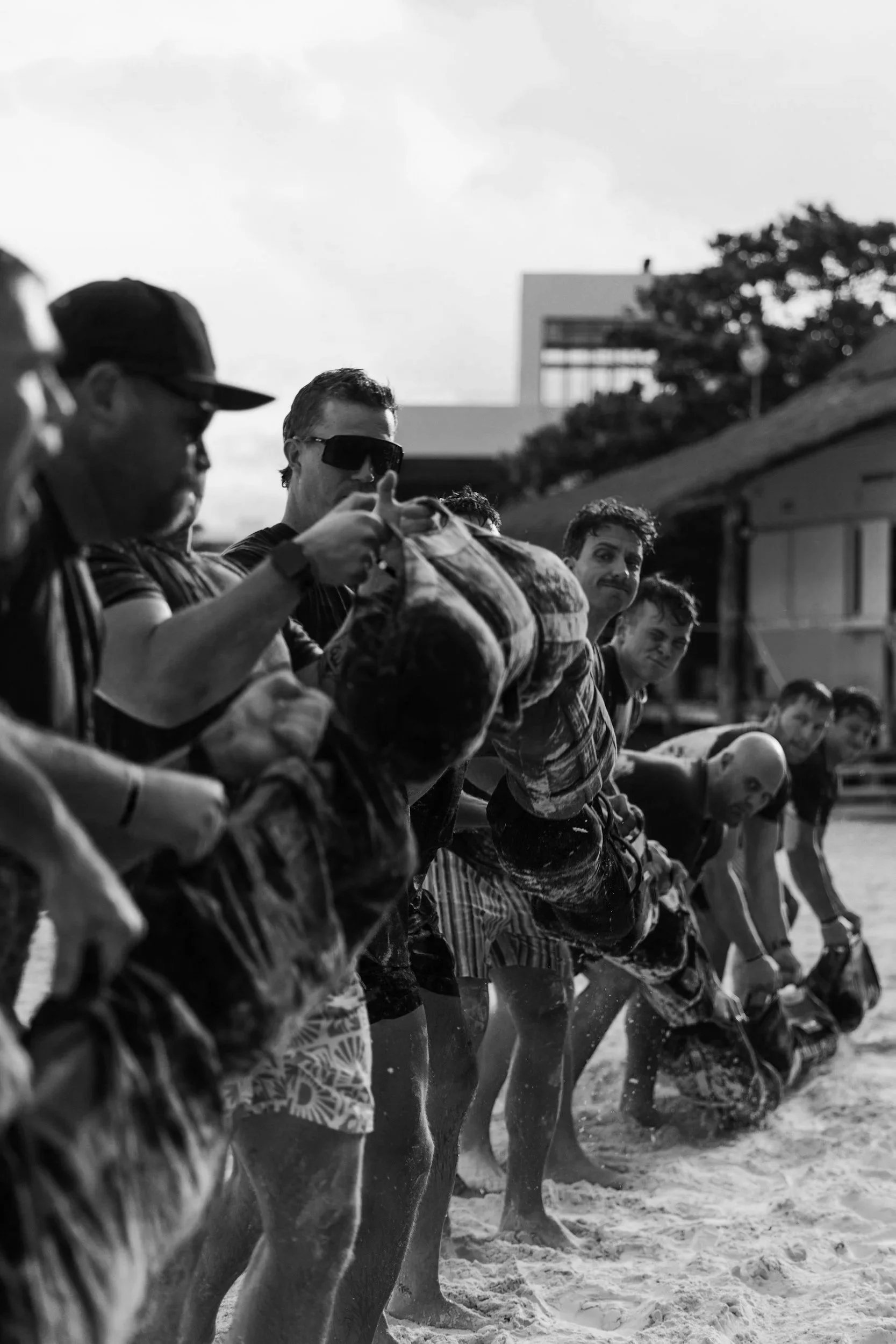 A black and white photo of several men standing in a line on the sandy beach, with some interacting with the water, in front of a building and trees.