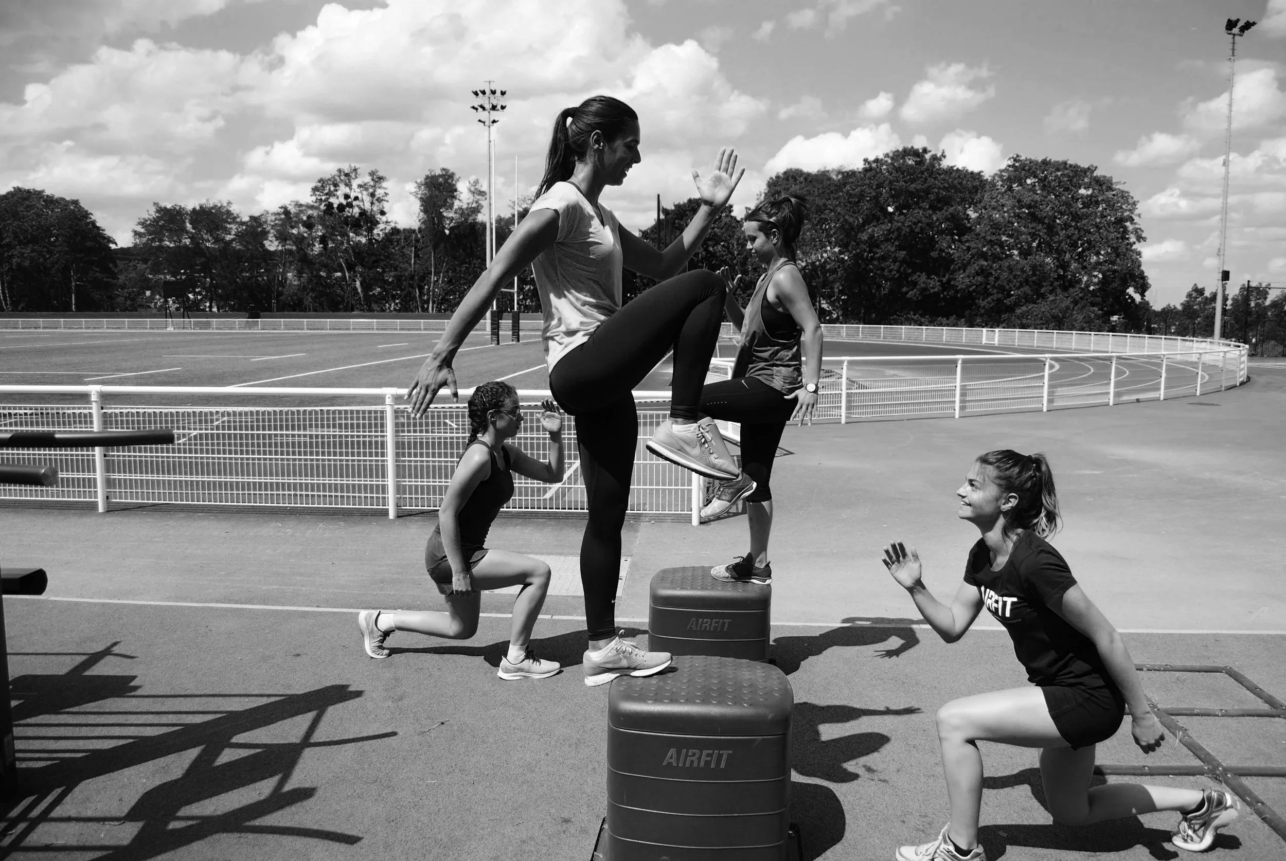 A group of women and girls practicing acro yoga outdoors on a sports field, with one woman balancing on a step while others support her from below, some kneeling and some standing, under a partly cloudy sky.