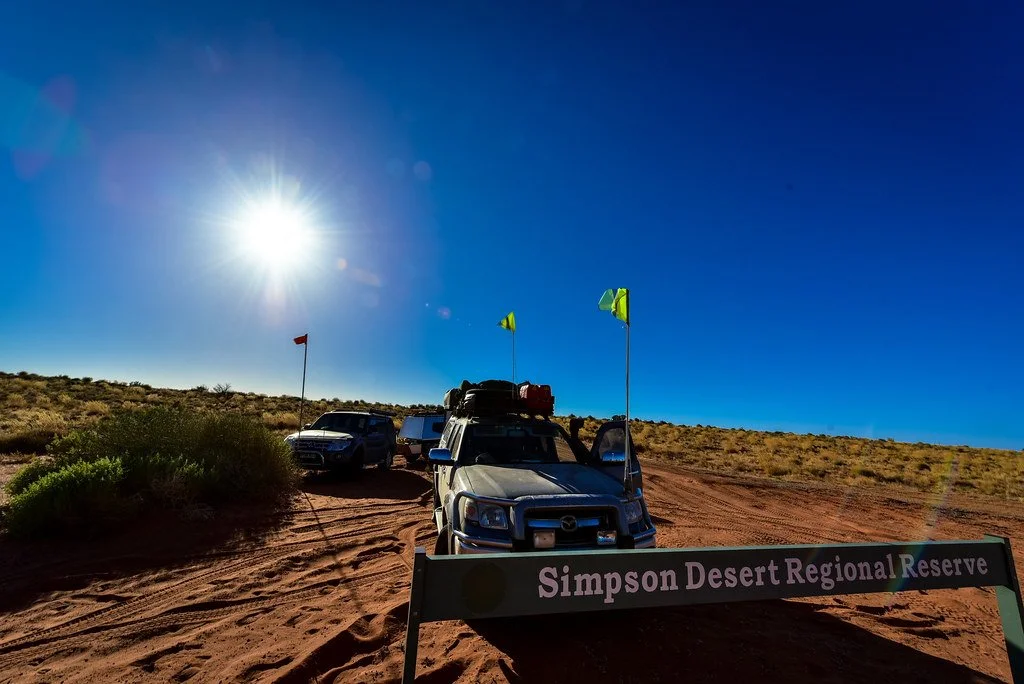 Vehicles parked on a dirt trail with a sign reading 'Simpson Desert Regional Reserve' and desert vegetation under a bright, sunny sky.