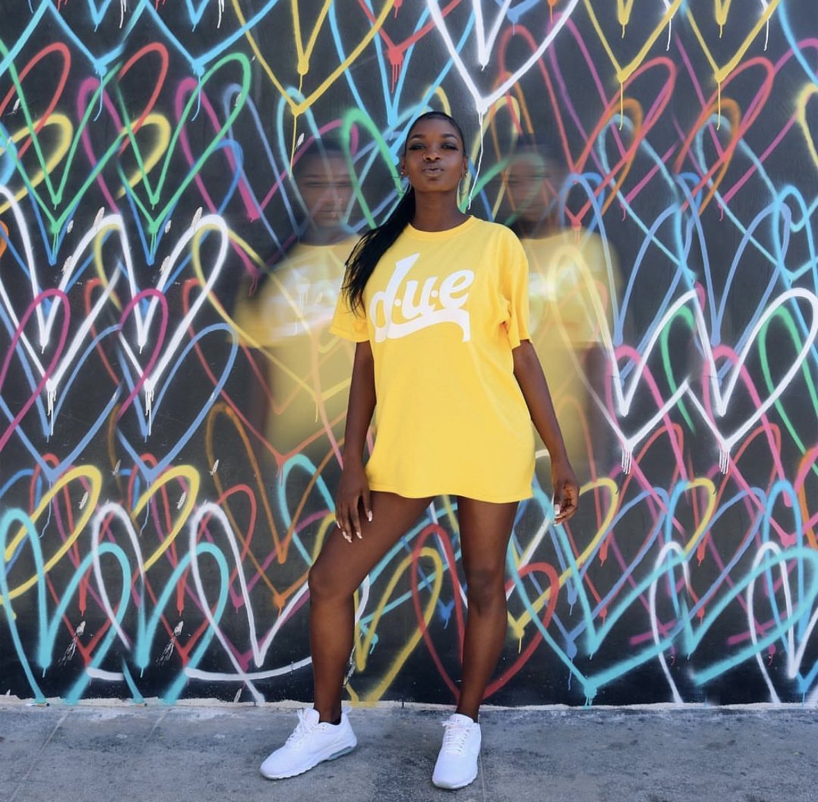 A woman wearing a yellow t-shirt with 'd.u.e' written on it, standing in front of a vibrant graffiti wall decorated with multicolored neon heart shapes.