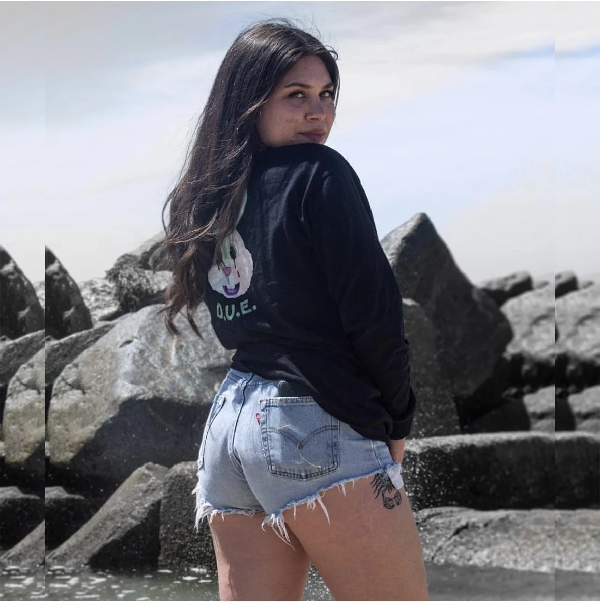 Young woman with long dark hair, wearing a black sweatshirt with a hamster logo and the text 'Q.U.E.', and light blue denim shorts with frayed edges, standing by large rocks or a jetty near water, looking over her shoulder at the camera, with a cloudy sky in the background.