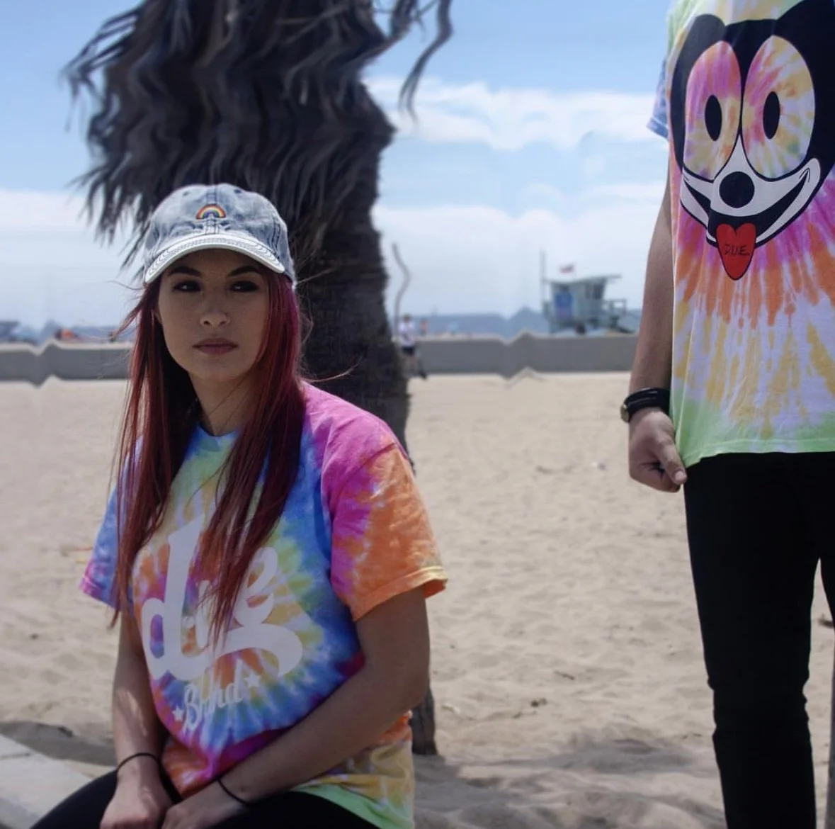 A young woman sitting on a beach, wearing a rainbow tie-dye t-shirt and a denim cap with a rainbow patch. She has long, reddish hair and is looking at the camera. A person standing next to her wears a similar tie-dye shirt with a Mickey Mouse face and the word "Love" on the tongue.