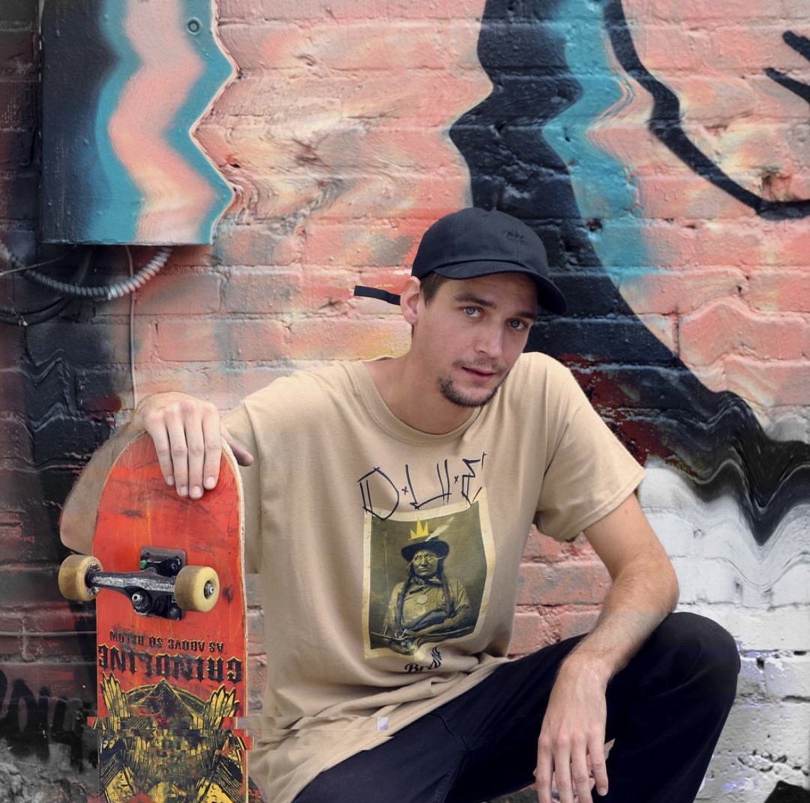 A young man with a beard and blue eyes, wearing a black cap and beige T-shirt with a graphic, sitting against a colorful street art mural on a brick wall, holding a skateboard with red and yellow graphics.