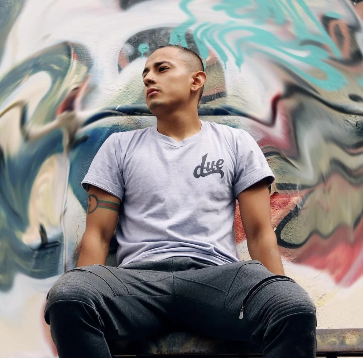 Young man sitting against a colorful graffiti wall, wearing a light gray t-shirt with 'dude' written on it and black pants, looking to the side.