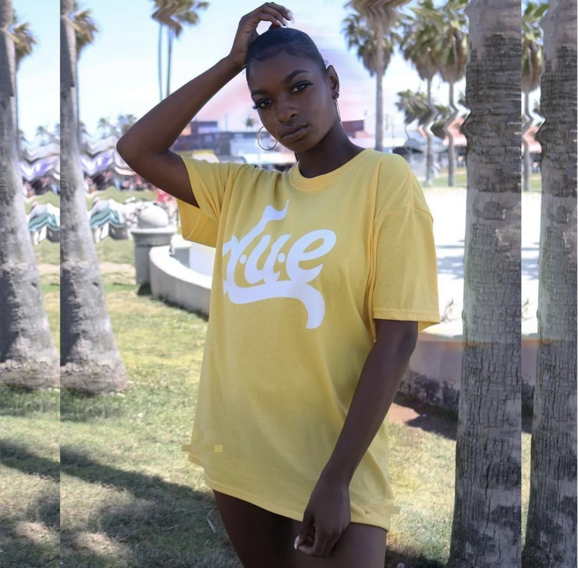 A young woman with dark skin and short hair standing outdoors near palm trees, wearing a yellow T-shirt with white lettering, looking at the camera.