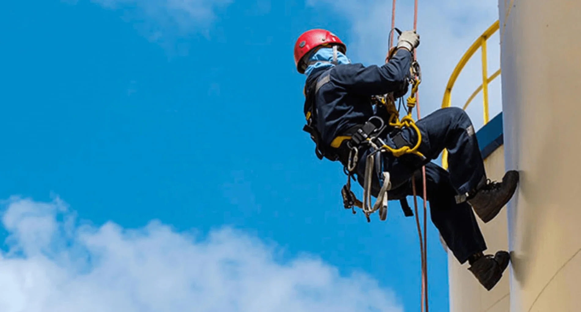 Trabajador en escalada industrial usando arnés y casco, asegurado con cuerda, mientras realiza mantenimiento en una estructura alta con cielo azul de fondo.