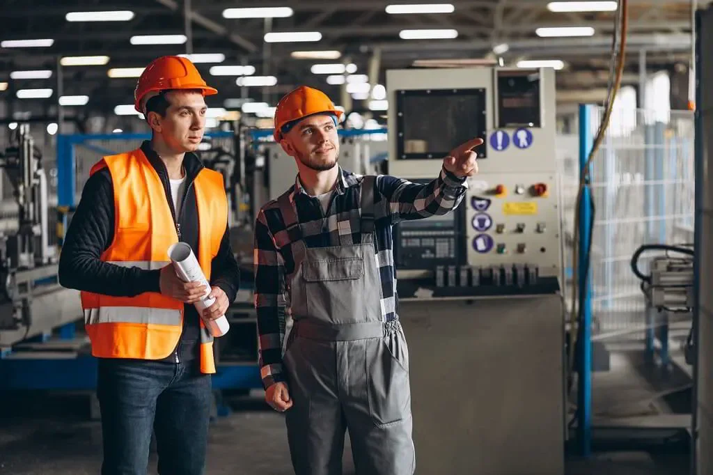 Dos trabajadores en una fábrica, uno con un casco y chaleco naranja, el otro con un casco y overol, señalando hacia un equipo de maquinaria industrial.