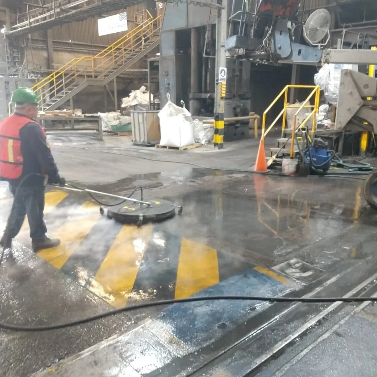 Persona limpiando el suelo en una fábrica o planta industrial, con maquinaria y escaleras en el fondo, usando equipo de protección como casco y chaleco de seguridad.