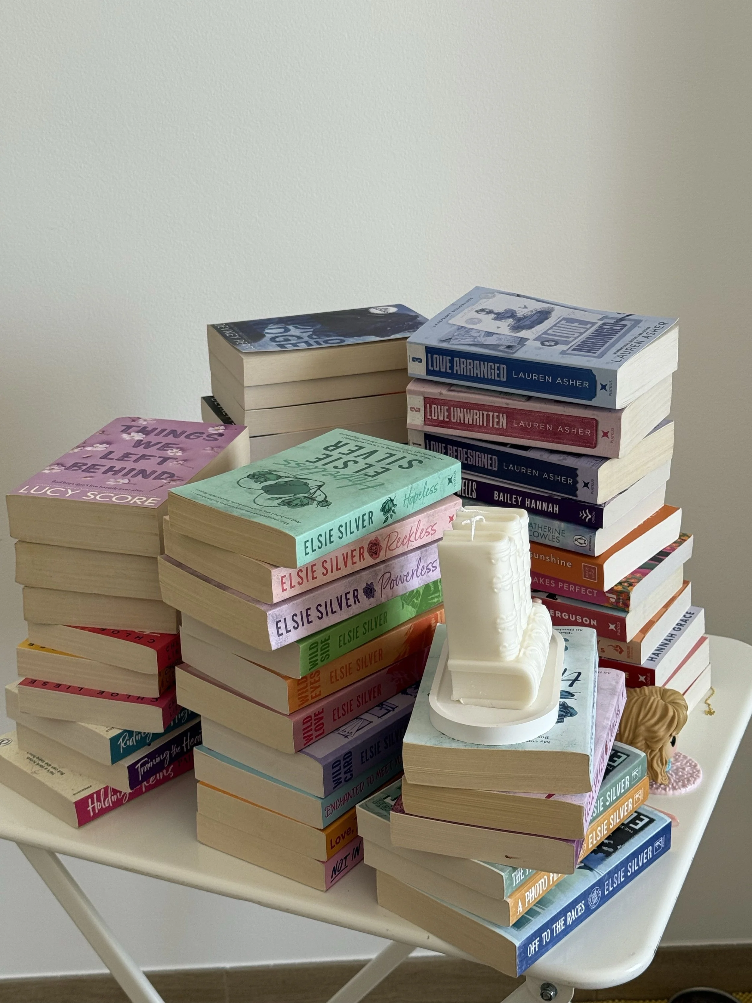 Several stacks of paperback books arranged on a white table next to a white decorative candle holder. The book covers are colorful, with titles including 'Holding Tears,' 'Hopeless,' and 'Off to the Races.' The background features a plain white wall.