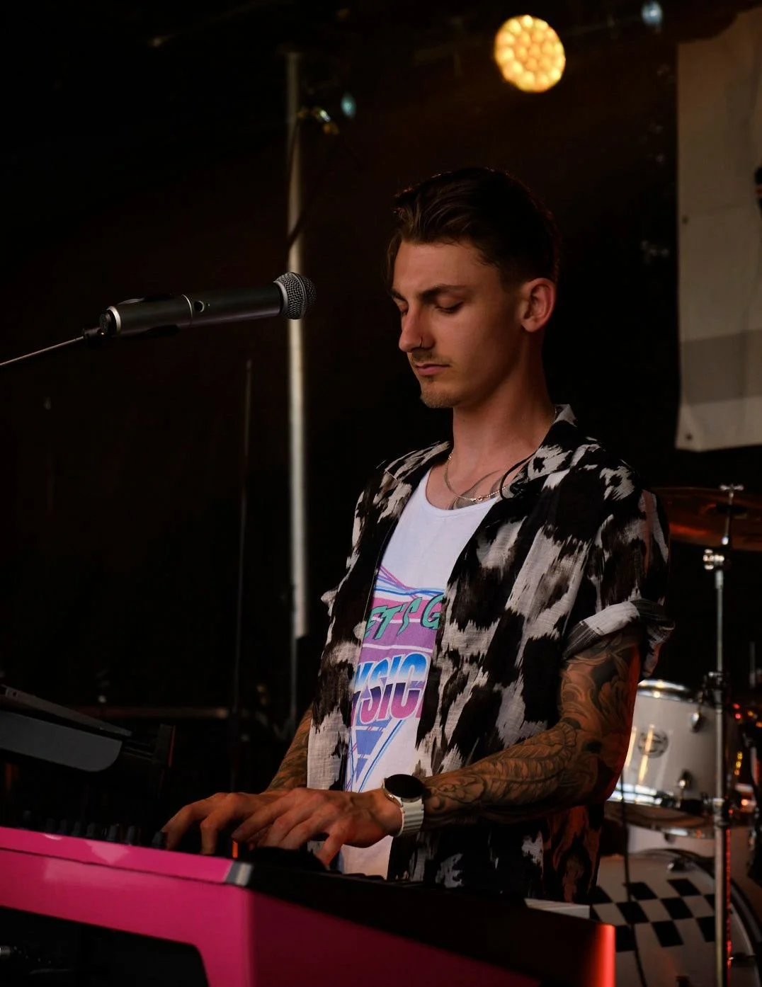 A young man with tattoos playing a pink keyboard on stage, wearing a patterned shirt and a graphic T-shirt, with a microphone in front of him and a drum set in the background.