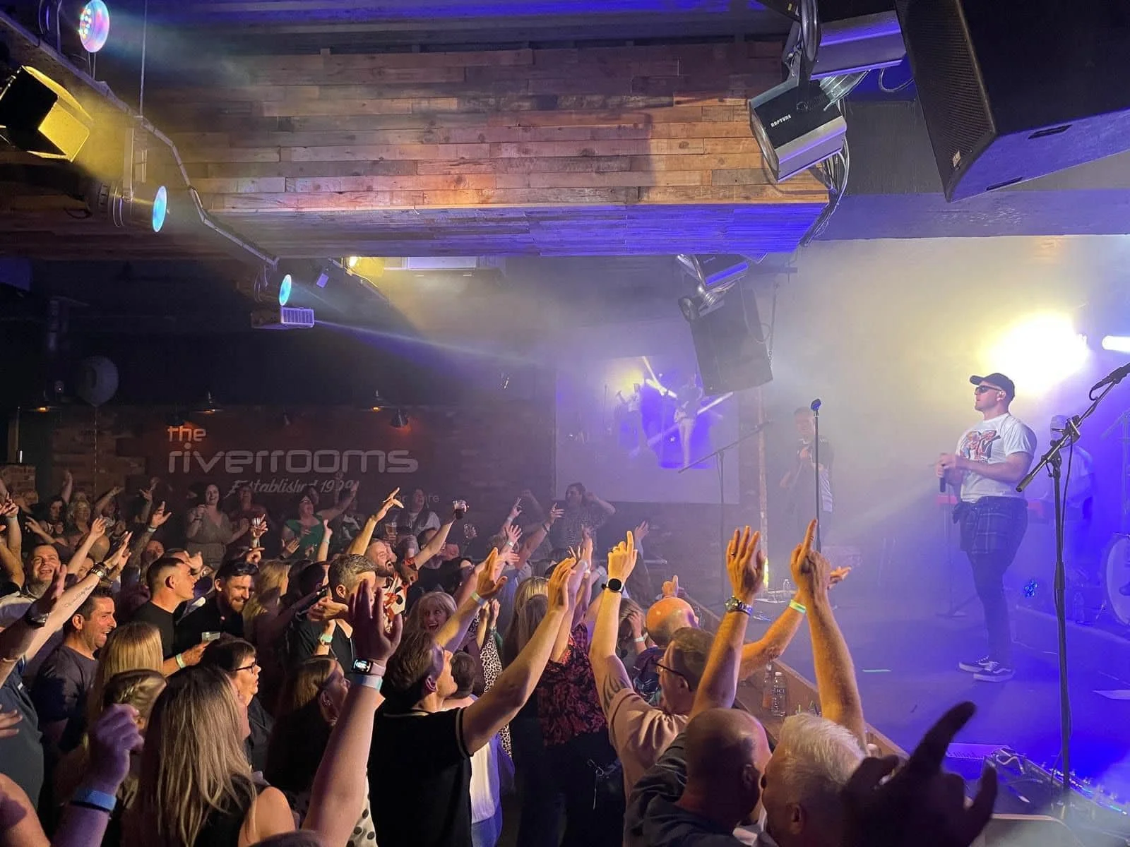 Concert scene at the Riverroom with a band performing on stage, audience cheering and raising their hands, stage lights shining brightly.