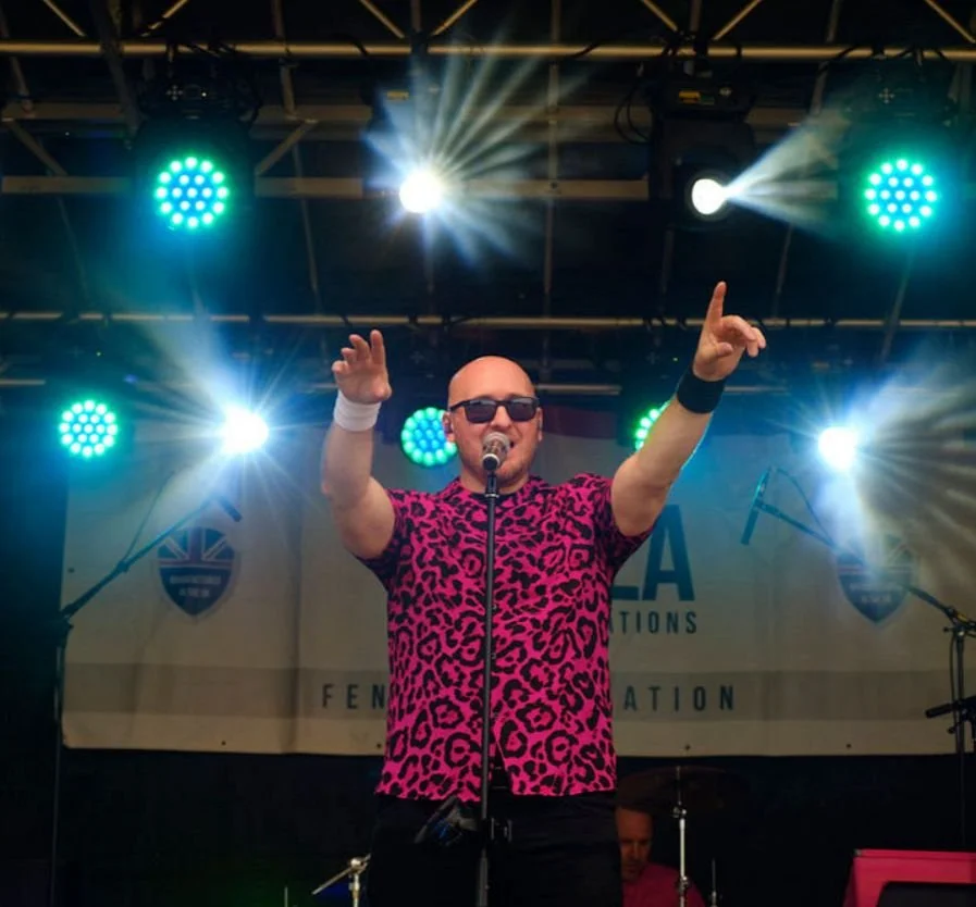 A man wearing sunglasses and a pink leopard-print shirt singing into a microphone on stage, with stage lights shining down on him.