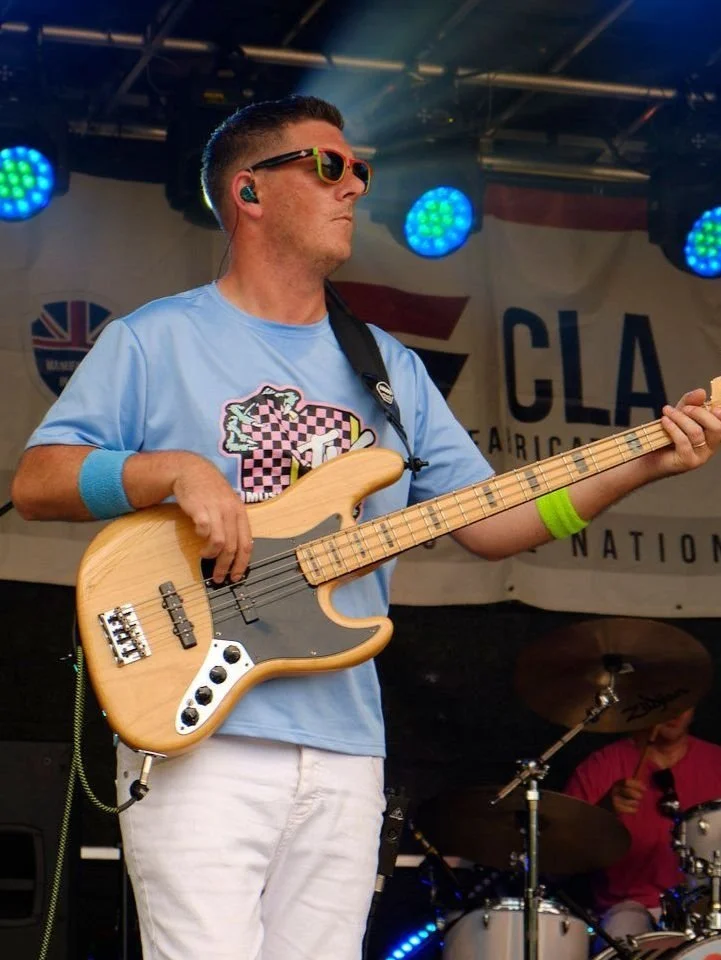 A man wearing glasses and colorful wristbands playing an electric bass guitar on stage with drums in the background.