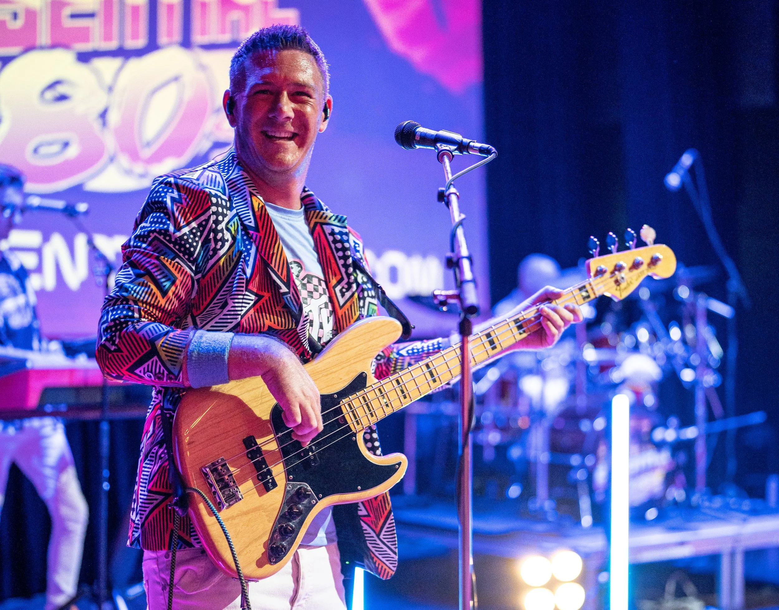 A man playing a bass guitar on stage during a live music performance, smiling at the audience with a colorful background and stage lights.