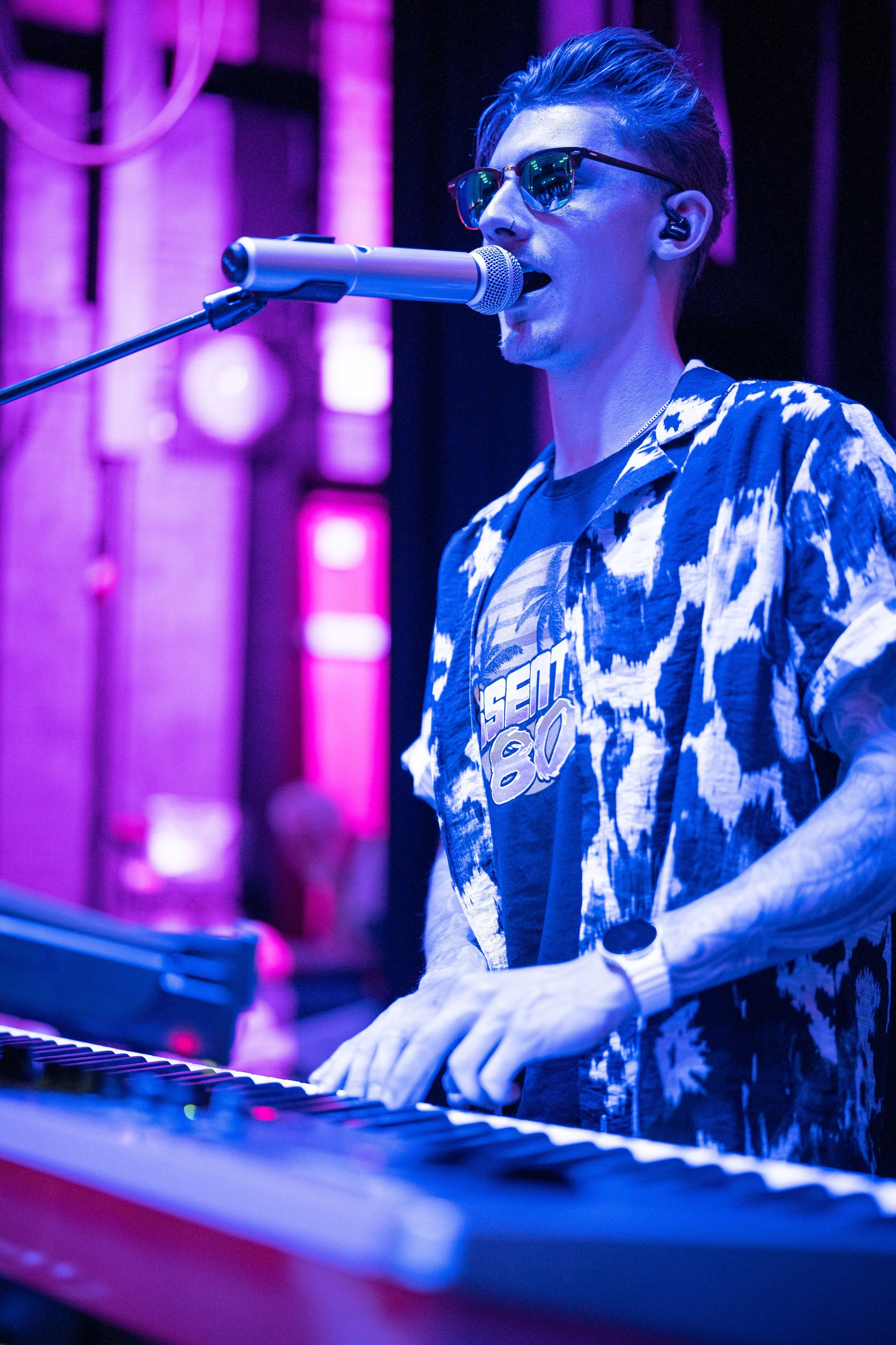 A young man wearing sunglasses and a patterned shirt is singing into a microphone while playing a keyboard in a dimly lit venue with purple and pink lighting.