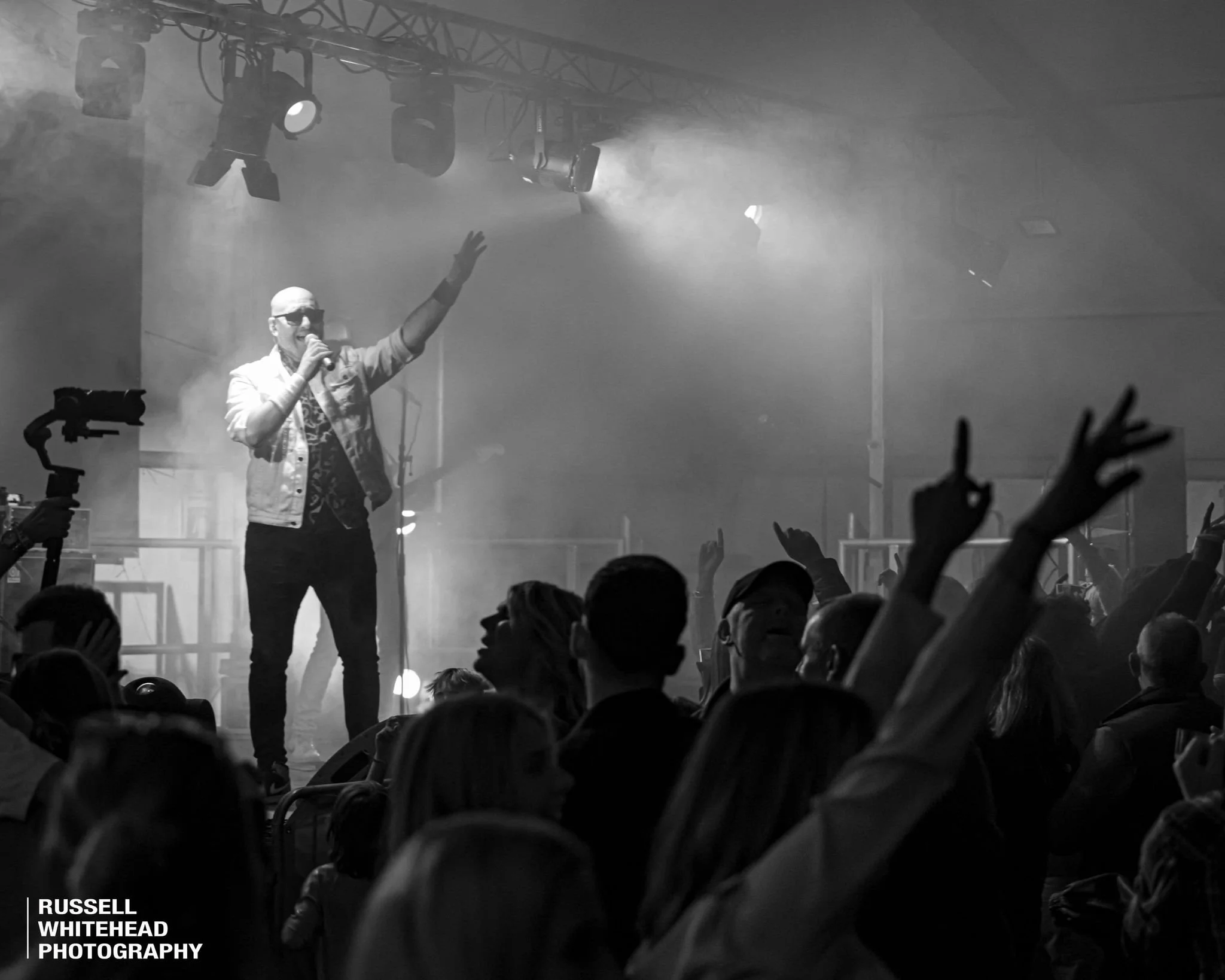 A black and white photograph of a performer on stage holding a microphone and raising one hand. The audience is visible in front, with several people raising their hands, some making the 'OK' gesture. Stage lighting is focused on the performer, with 