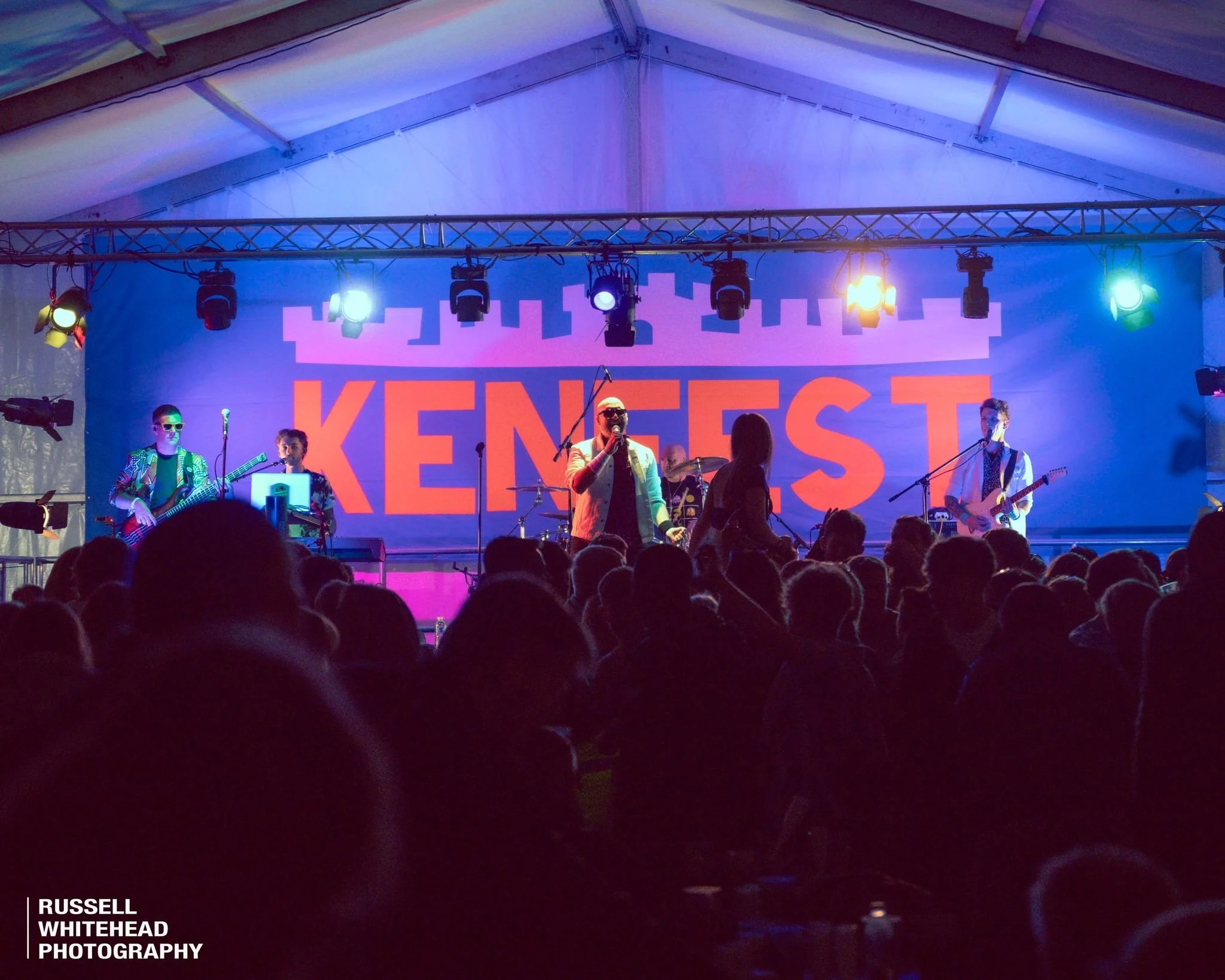 A band performs on stage at an event called 'KENGST' with a crowd in front. The stage is inside a tent with colorful lighting.