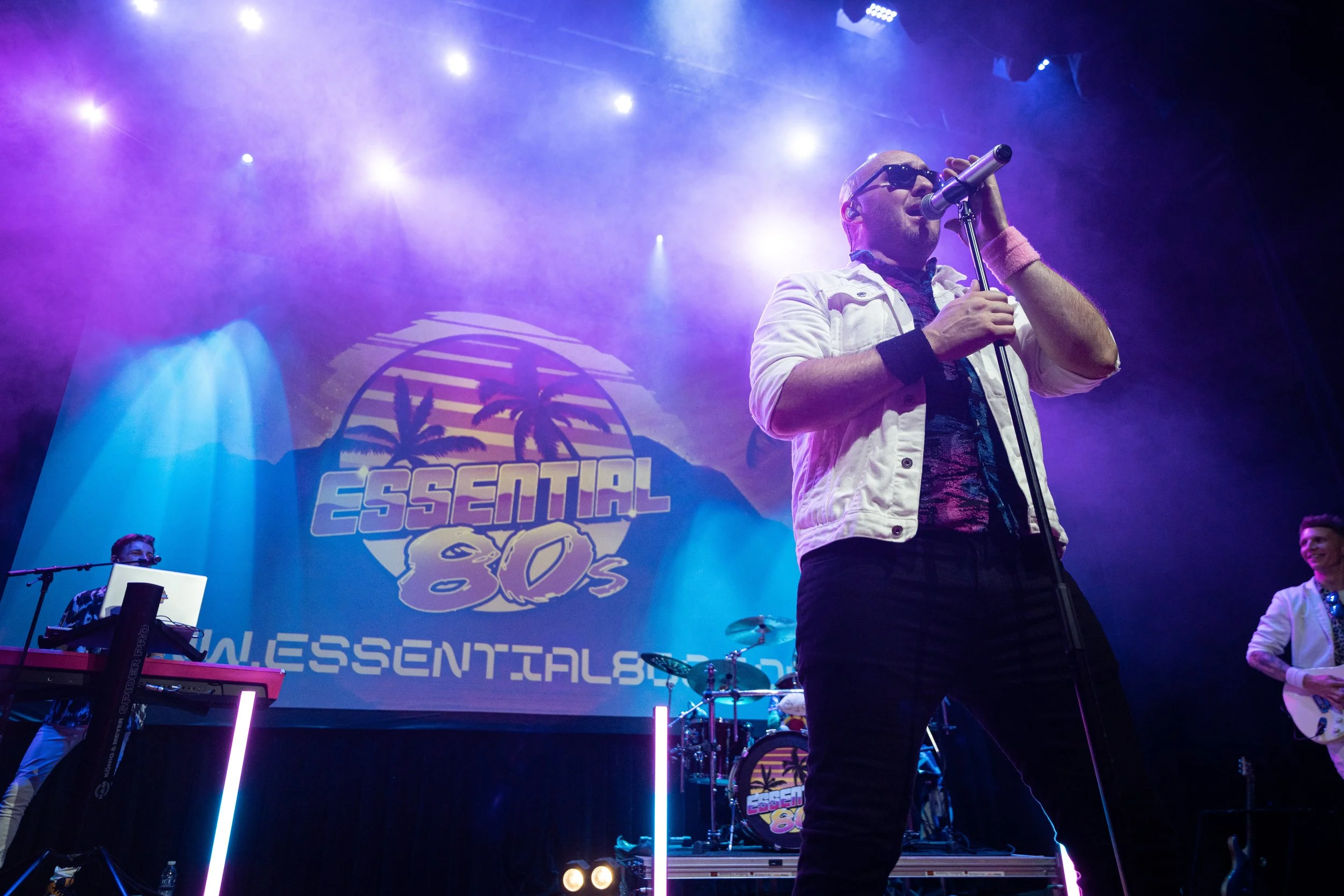 A band performing on stage with a large screen in the background displaying the words 'Essential 80s' with a sunset and palm trees graphic.