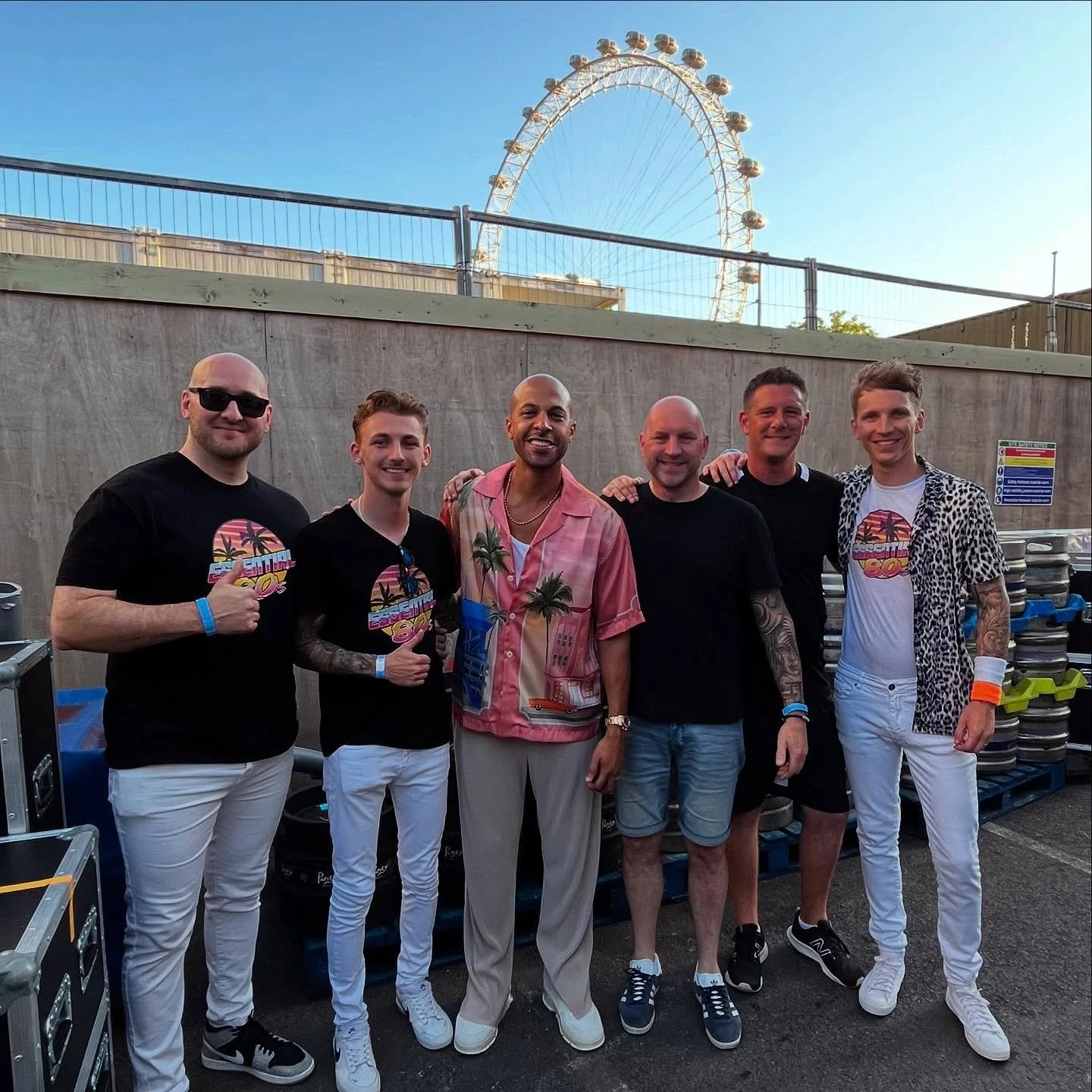 Six men standing together outdoors with a Ferris wheel in the background, all smiling and posing for the photo.