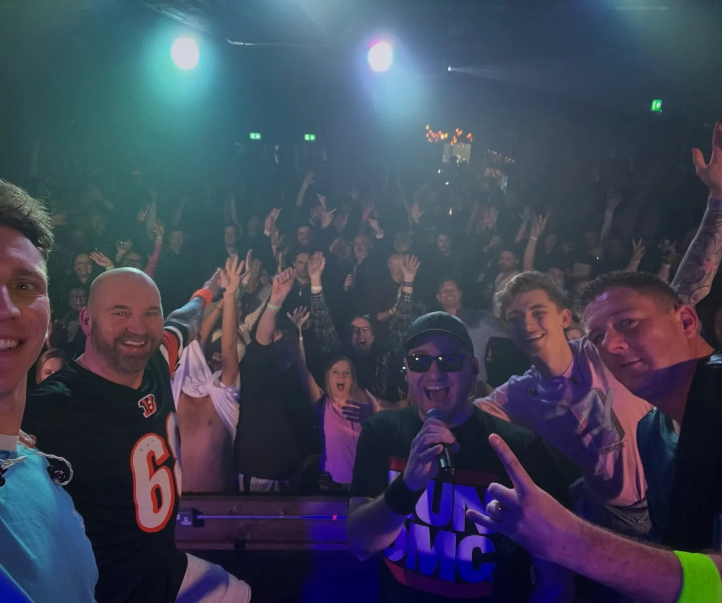 Group of people at a concert or event, smiling and posing for a selfie with a large crowd in the background, colorful lighting overhead.