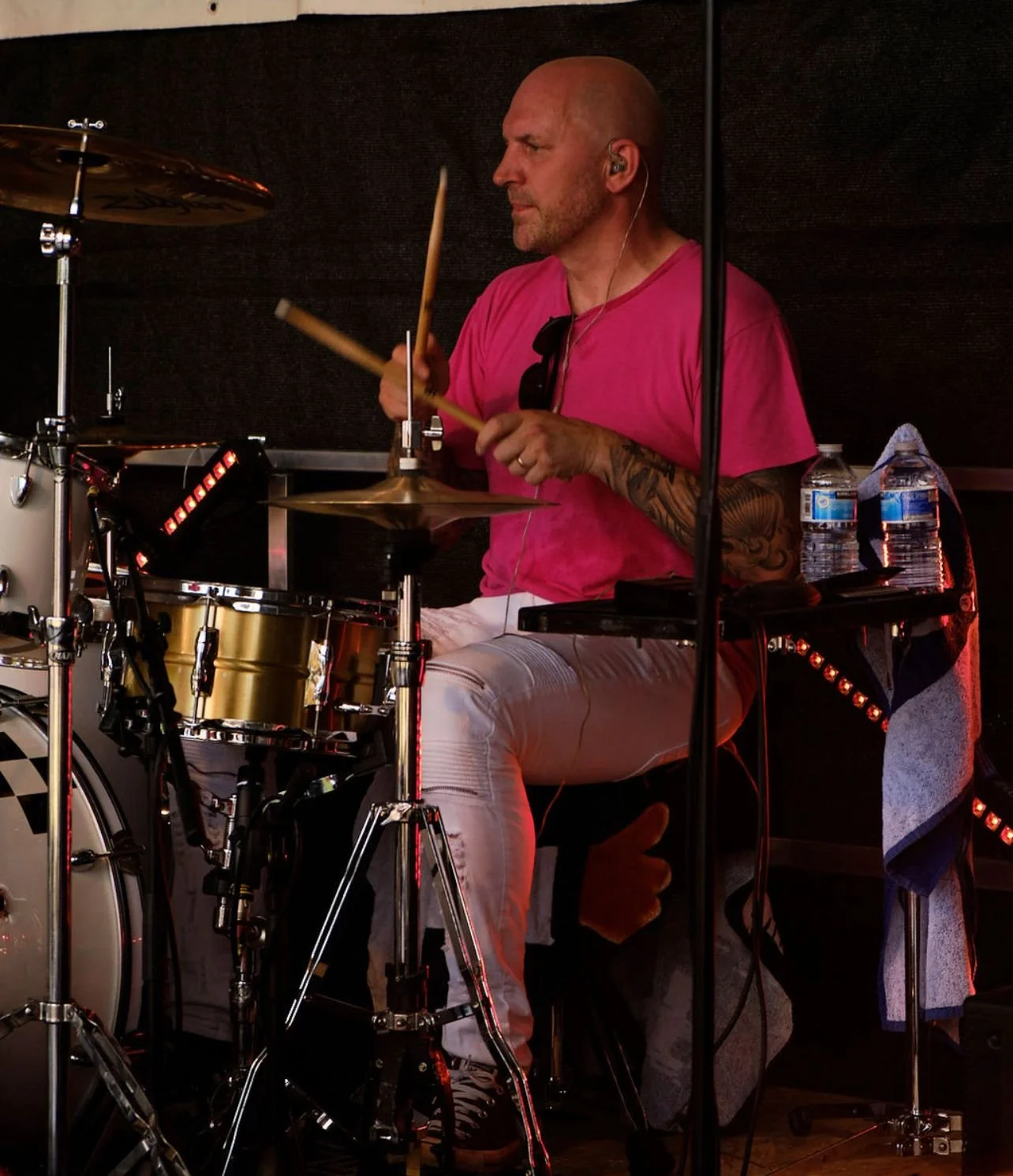 Bald man playing drums on stage, wearing a pink T-shirt, white pants, and black sneakers, with water bottles and a towel on a table nearby.