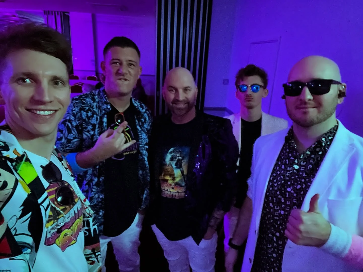 Group of five men at a party or club, wearing colorful and stylish clothing, some with sunglasses, under purple lighting.