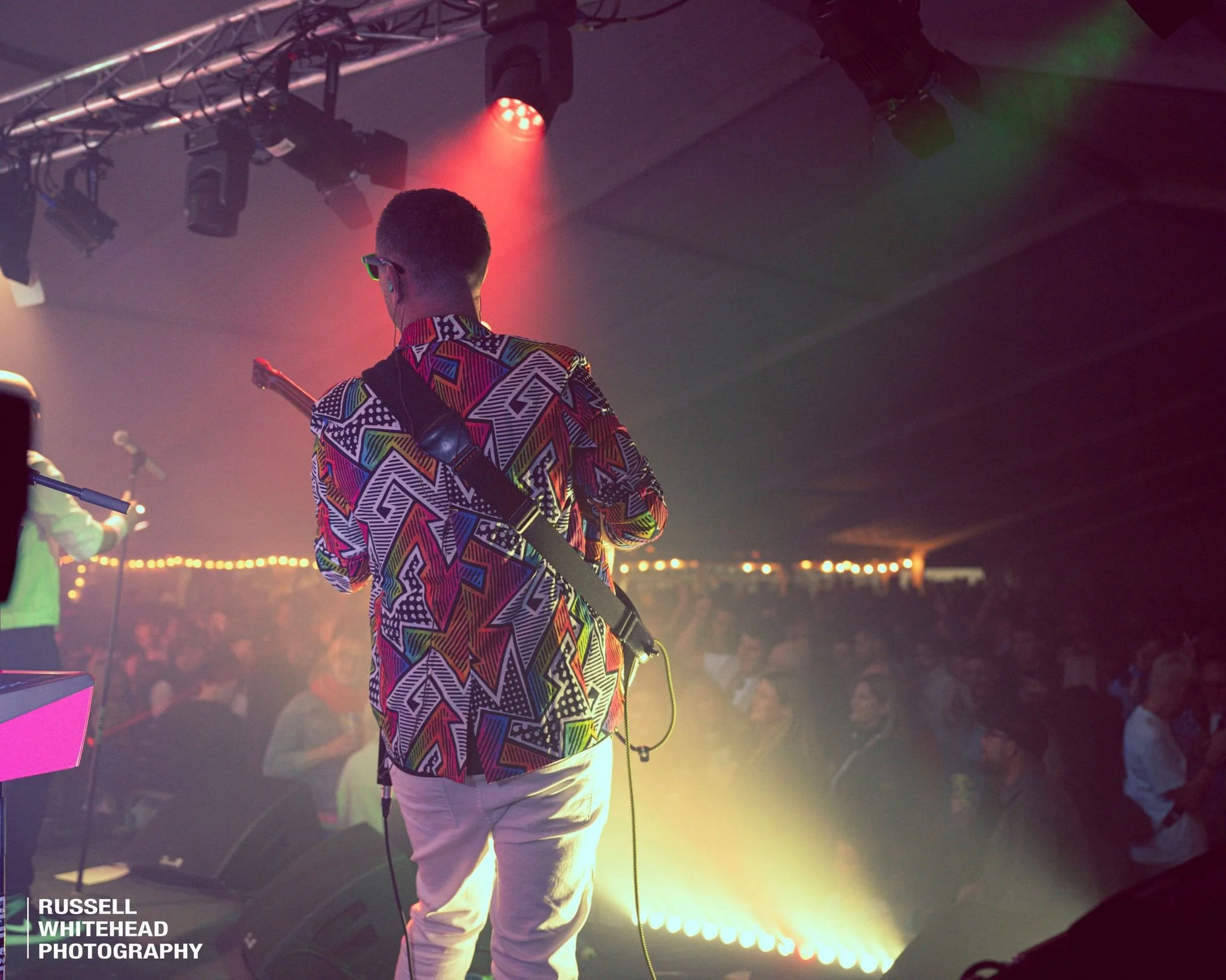 Musician on stage with colorful patterned shirt playing guitar, audience in background with stage lighting and fog.