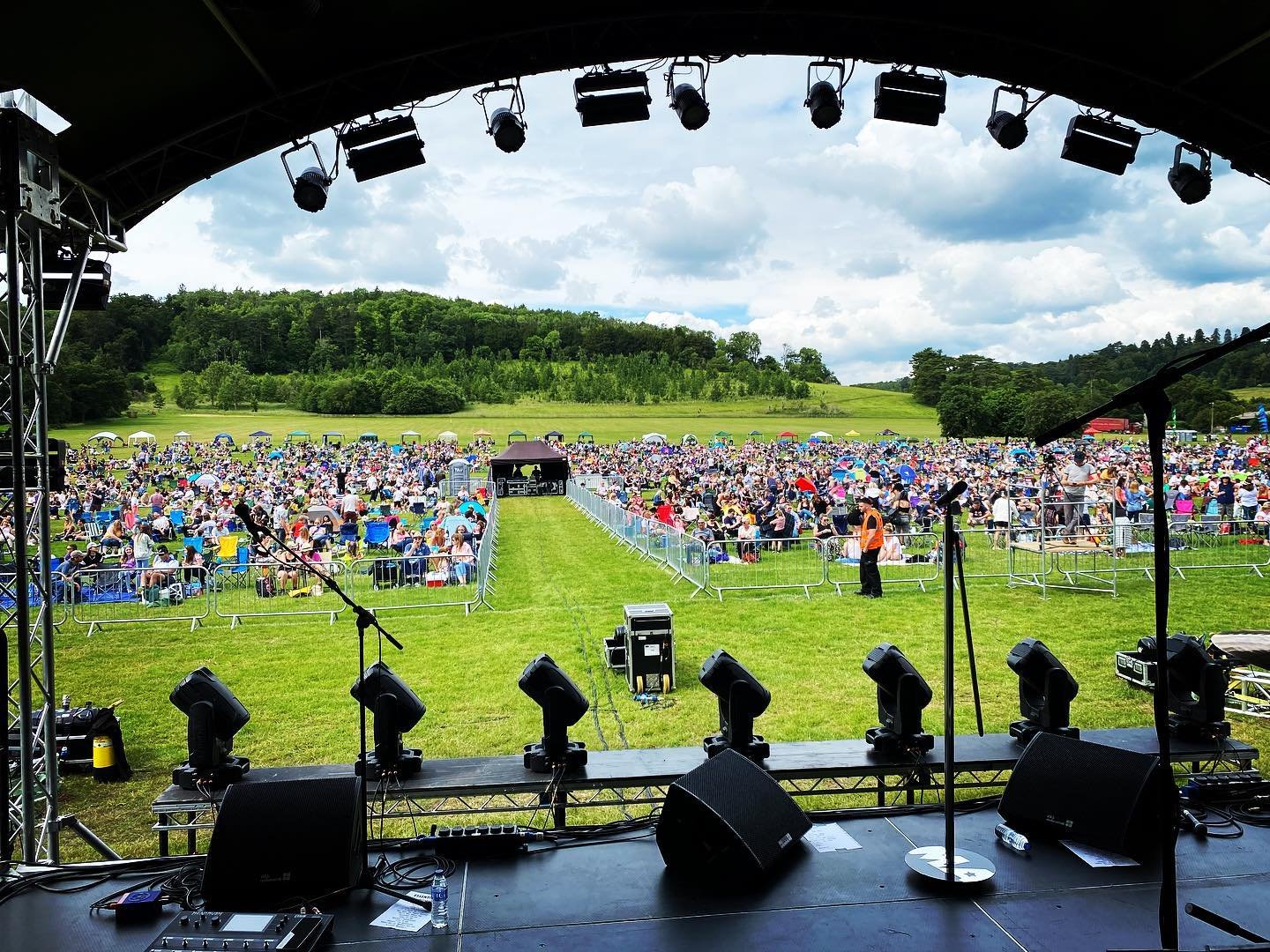 View from a stage overlooking a large outdoor concert with a crowd sitting on grass and picnic chairs, surrounded by green hills under a partly cloudy sky.