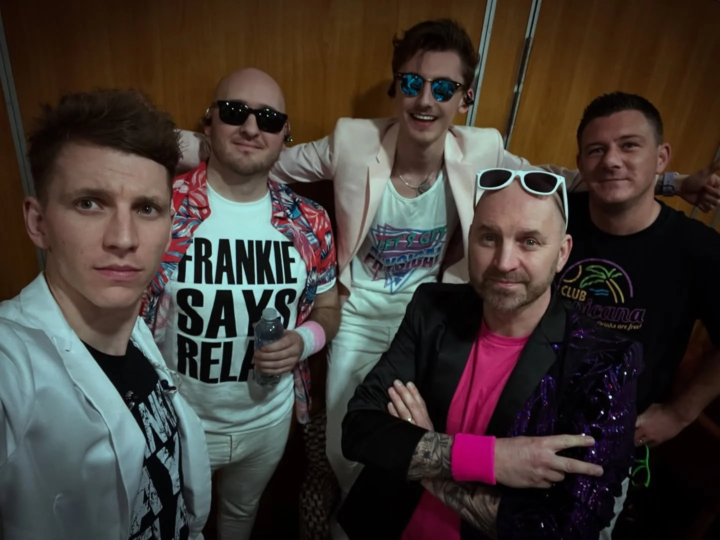 Group of six men posing together, some wearing sunglasses and casual colorful clothing, in an indoor setting with wood-paneled walls.